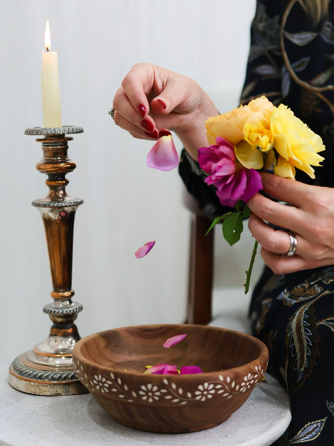Person placing pink flower petals into a wooden bowl with yellow and pink flowers in hand, beside a lit candle on a silver candlestick.