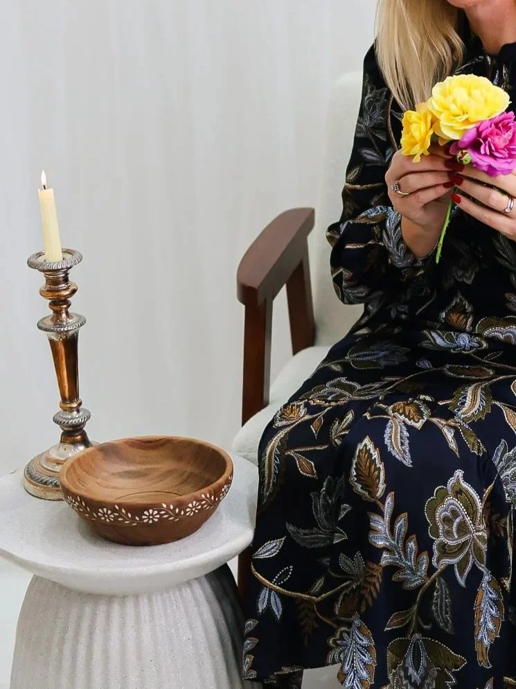A woman holding a bouquet of yellow and pink flowers sitting next to a side table with a lit candle on a candlestick and a wooden bowl.