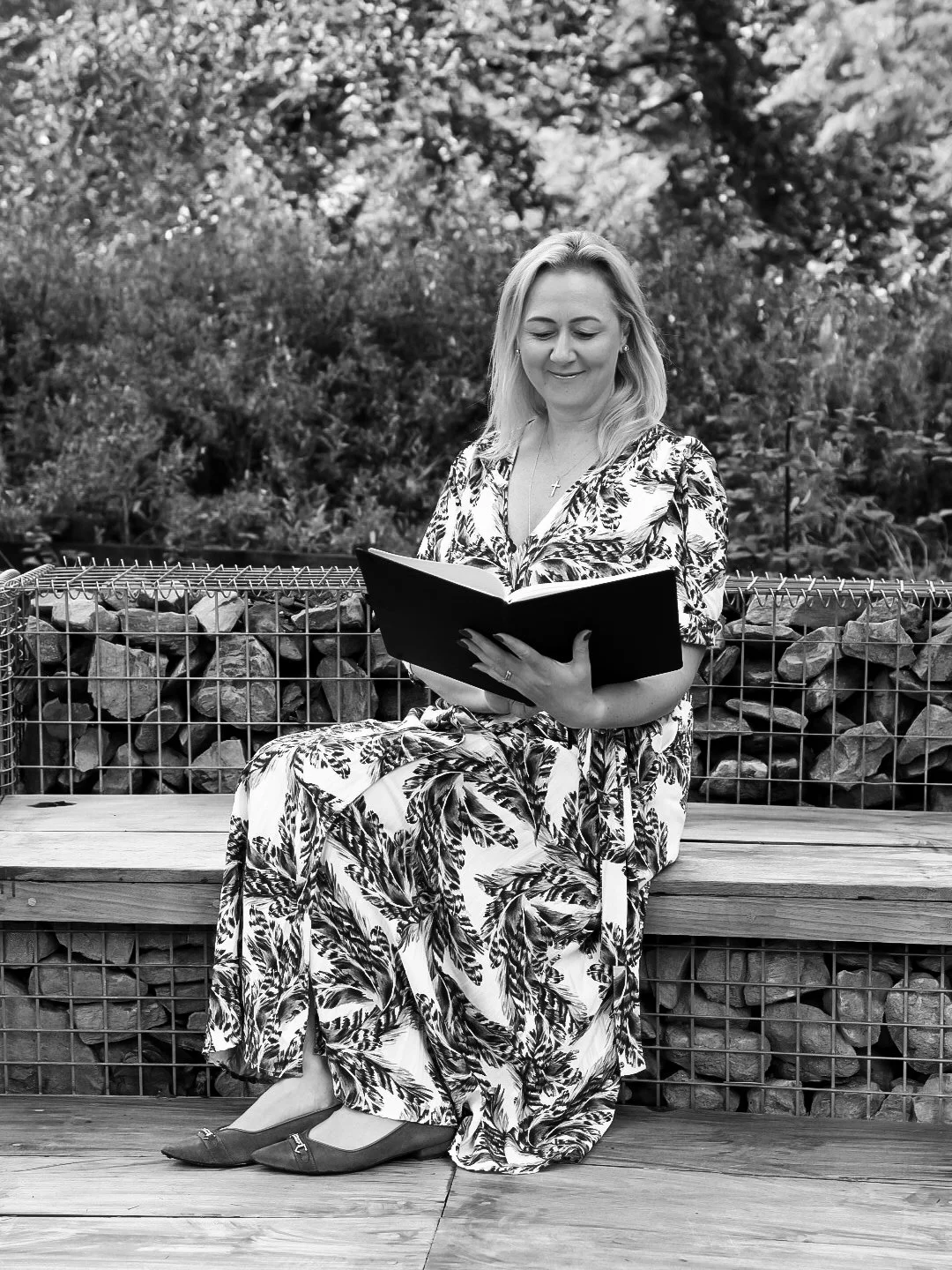A woman in a floral dress sits on a wooden bench outdoors, reading a book, with a backdrop of trees and a metal fence.