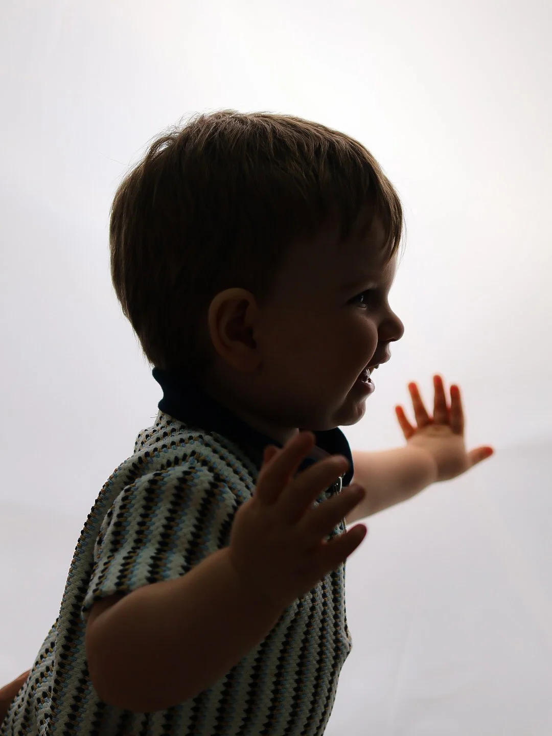Side profile of a young boy with light brown hair reaching out with both hands against a plain white background.
