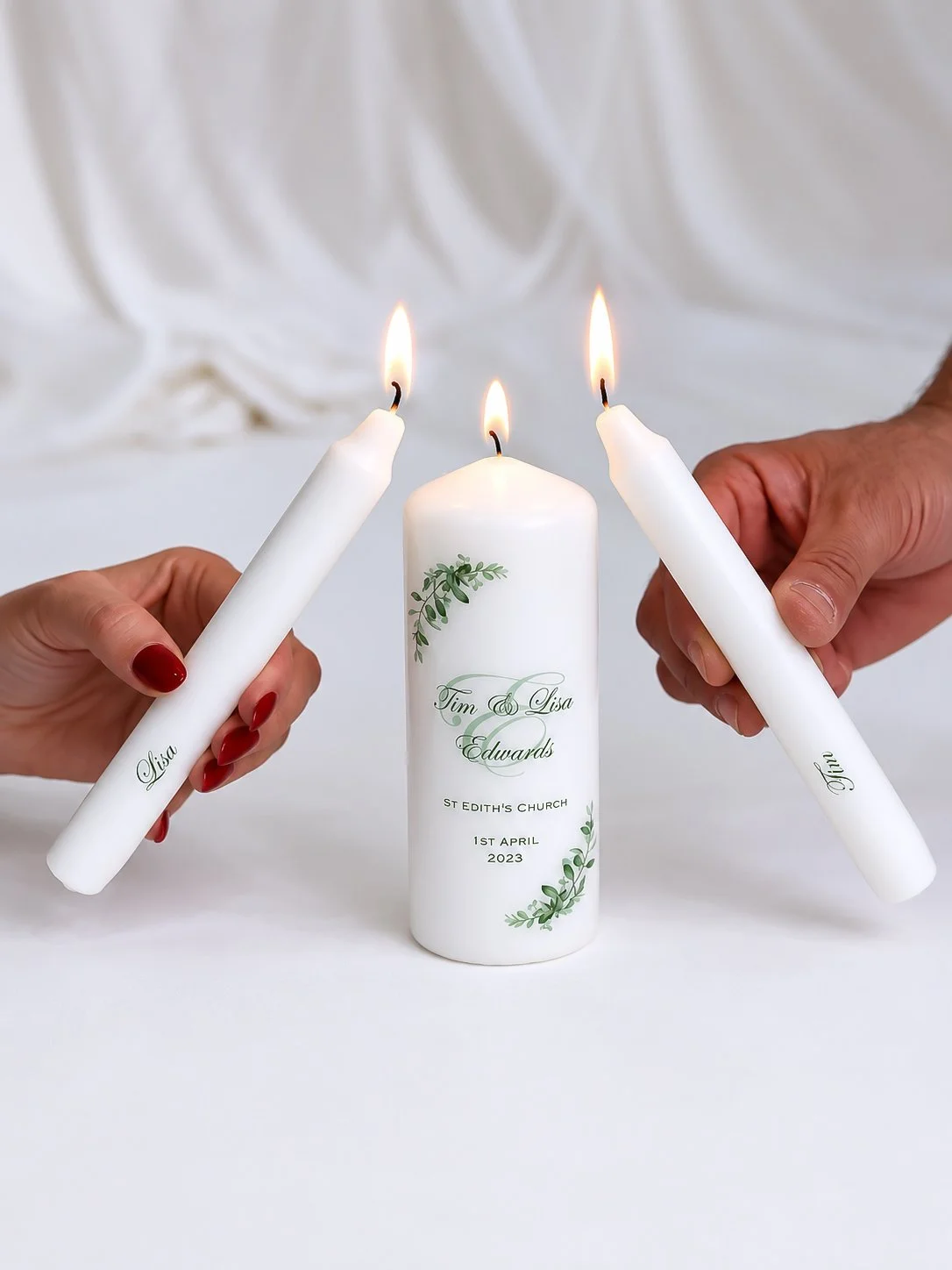 Two hands holding white candles with flames, around a larger white candle with personalized text and a green leaf design, on a white surface.