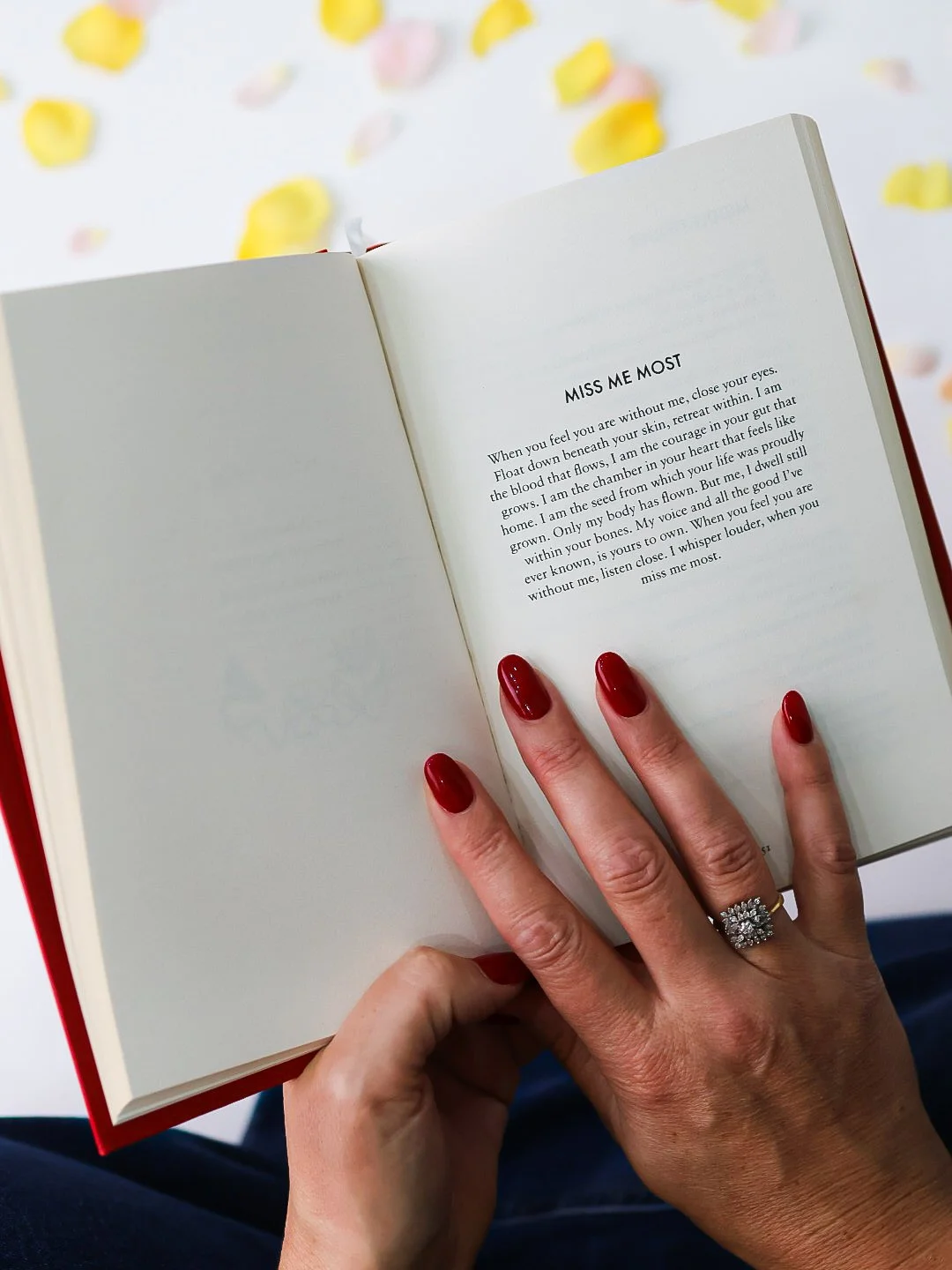 A person with red painted nails and a diamond ring is holding open a book with a poem titled 'Miss Me Most.' The background has yellow and pink flower petals scattered on a white surface.