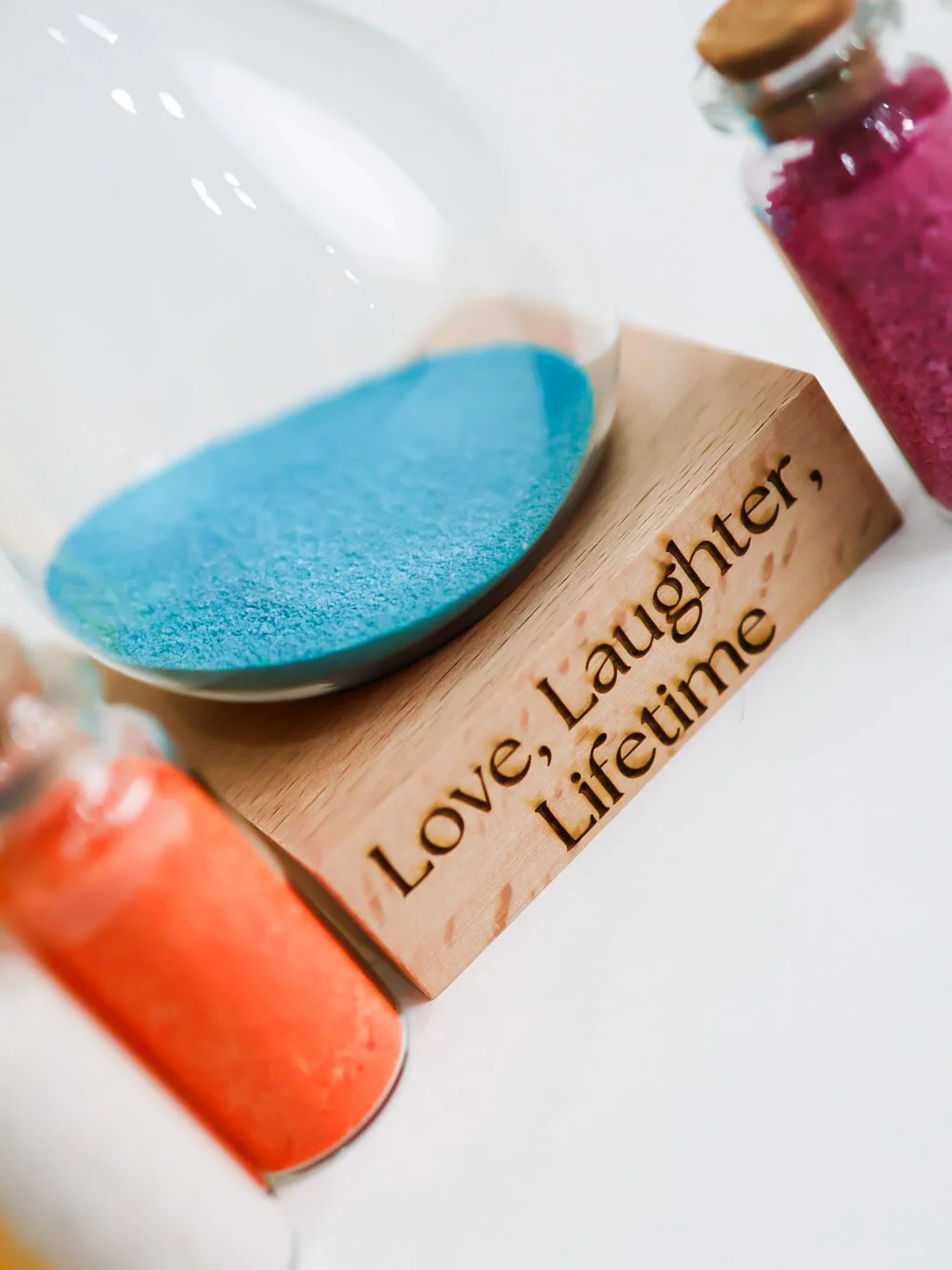 A glass container with blue powder, a small jar with pink glitter, and a small jar with orange glitter, on a wooden stand engraved with the words 'Love, Laughter, Lifetine'.