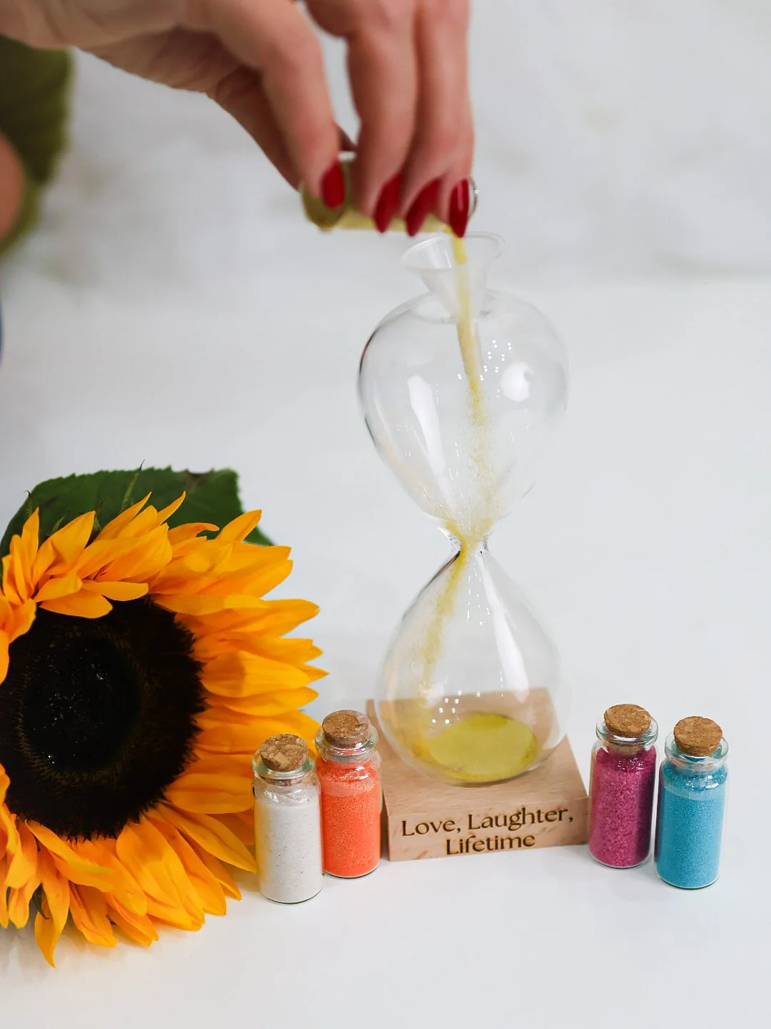 A hand with red painted nails pouring yellow sand into an hourglass, with a sunflower and four small jars of colorful sand nearby, on a white surface with a wooden block that reads "Love, Laughter, Lifetime."