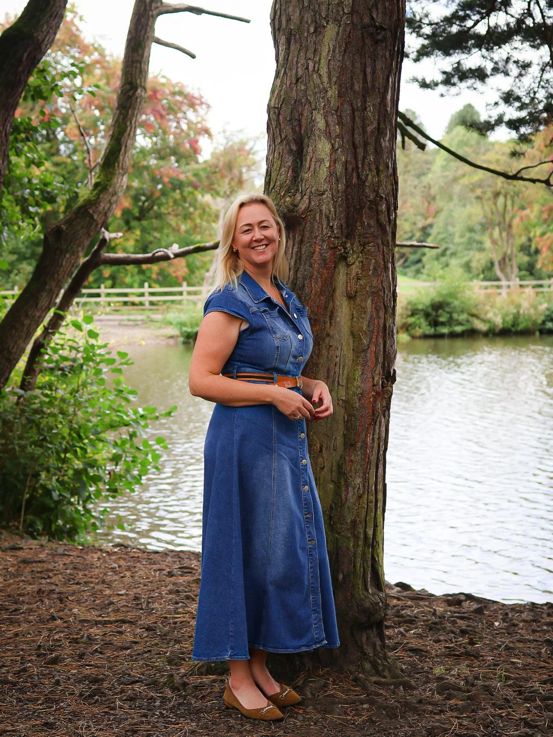 A woman in a denim dress standing by a large tree near a body of water in a park.