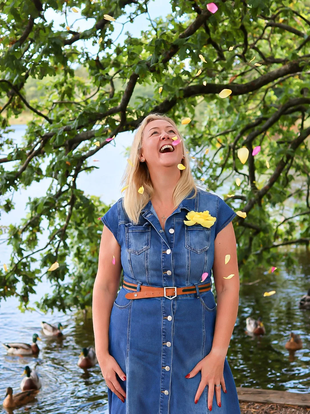 A woman in a denim dress with a belt, standing outdoors near a lake, surrounded by ducks and dropping flower petals from a tree above, smiling and looking up.