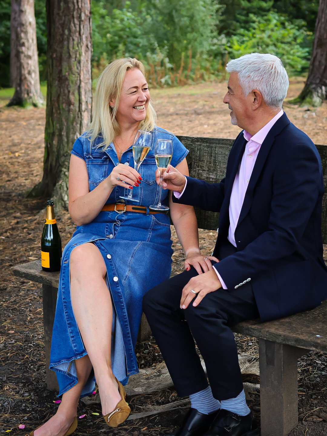 A woman and a man sitting on a bench in a forest, smiling and clinking glasses of champagne.