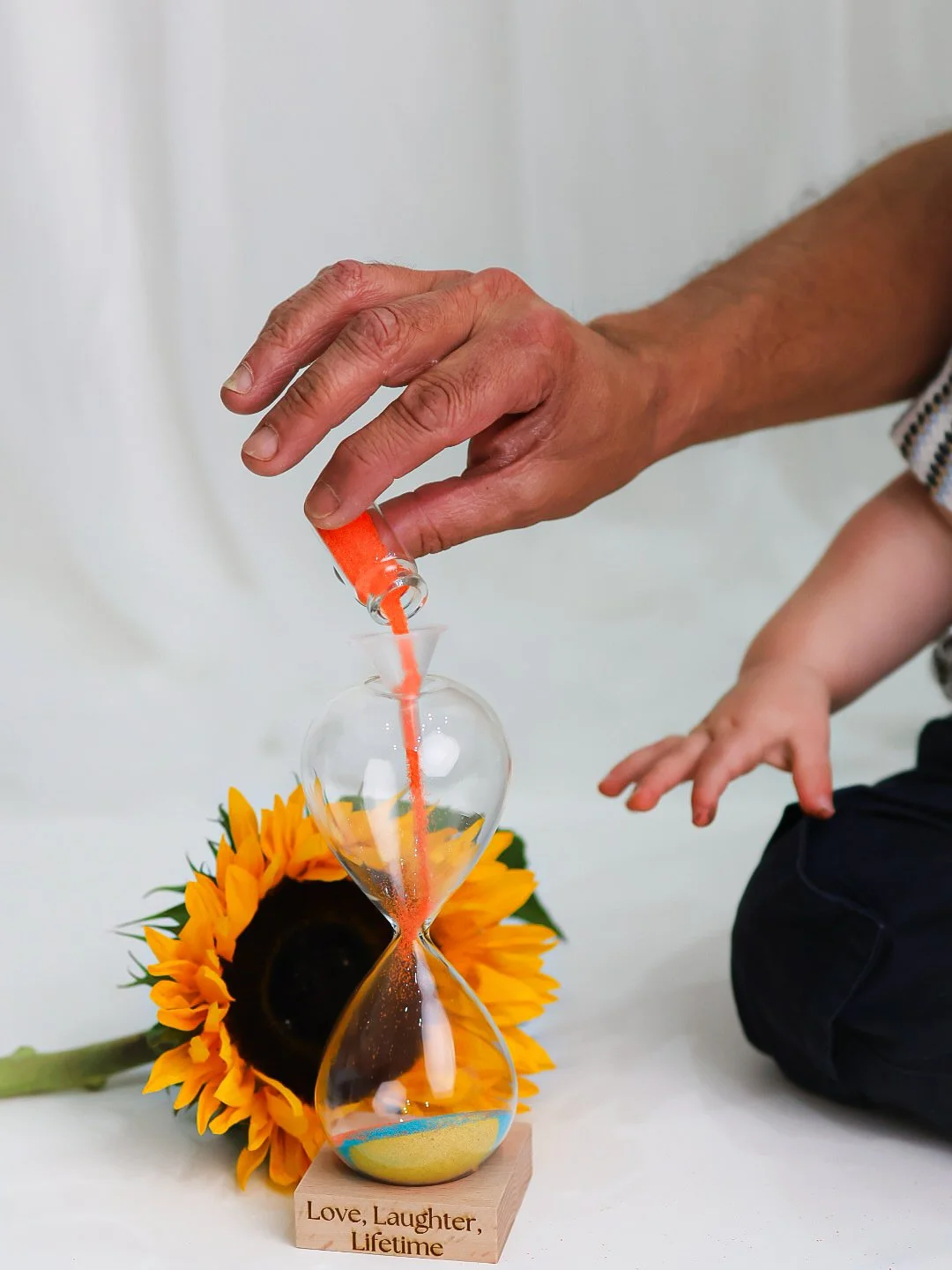 Man pouring orange sand into an hourglass with a sunflower in the background and a wooden base engraved with 'Love, Laughter, Lifetime'.