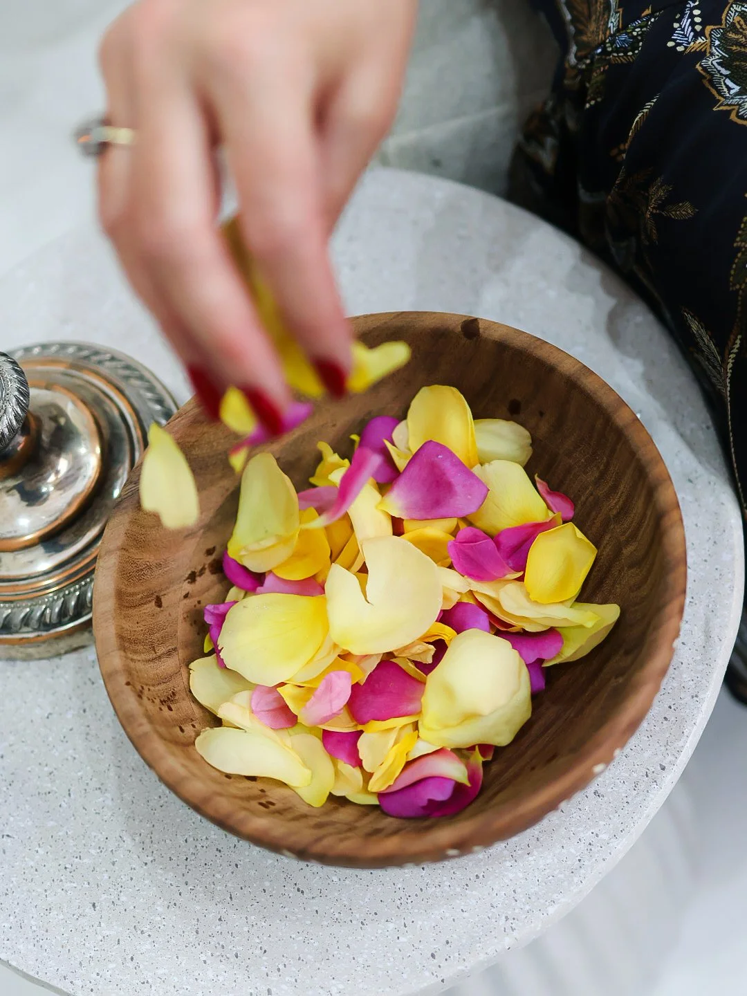 A person placing yellow and pink rose petals into a wooden bowl on a gray table.