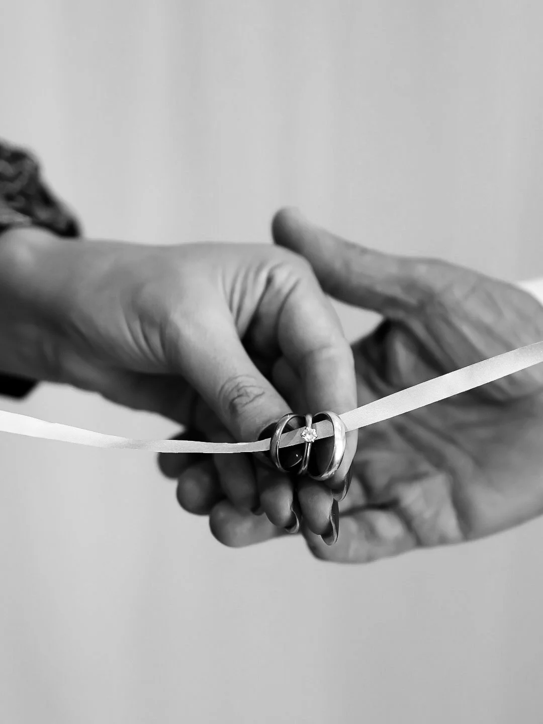 Close-up black-and-white photograph of two hands holding wedding bands with a small diamond, symbolizing marriage, with a plain background.