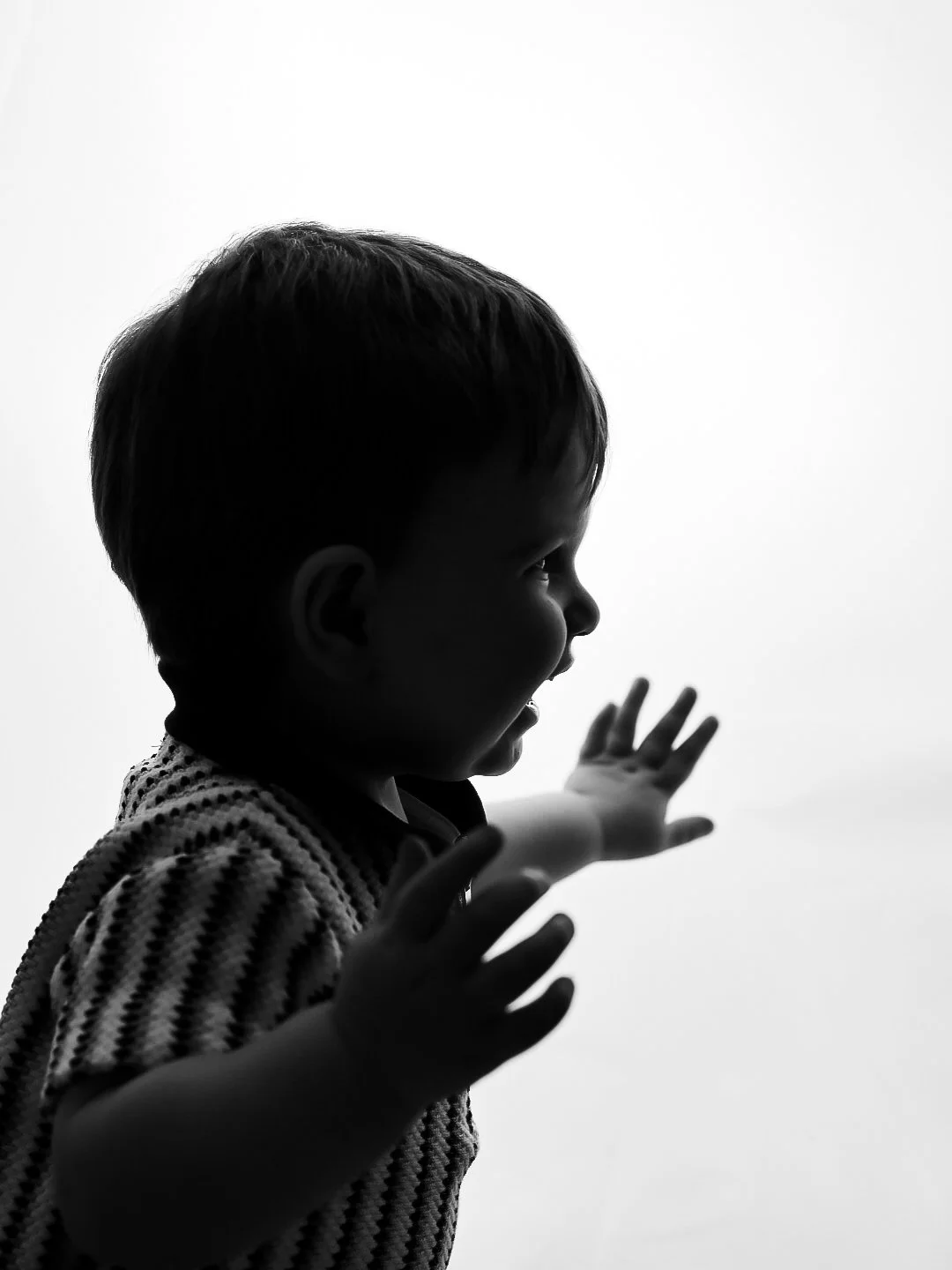 Silhouette of a young child with short hair, reaching out with both hands, in front of a plain, bright background.