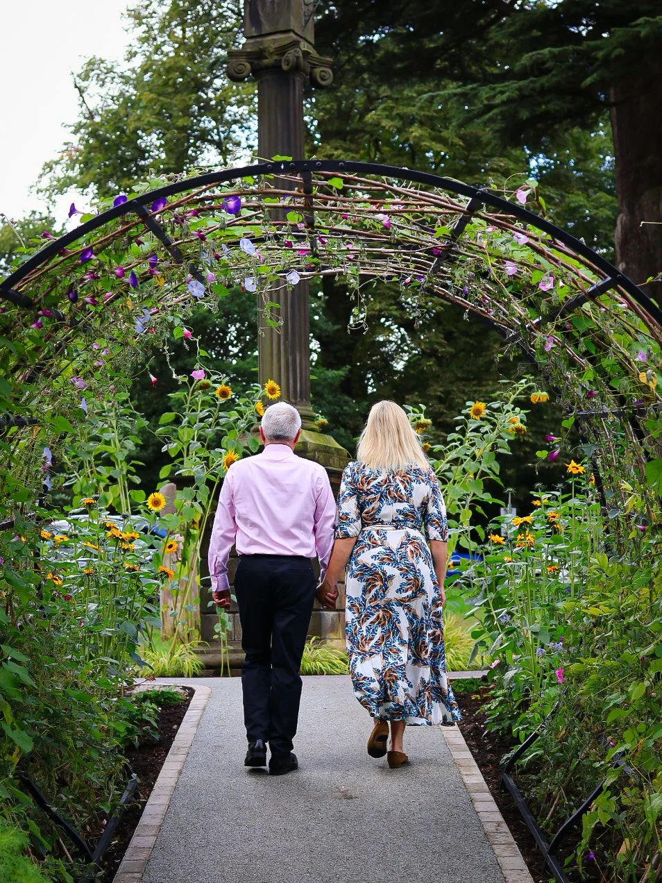 An older man and woman walking hand in hand through a garden archway decorated with flowers, with sunflowers and greenery around them.