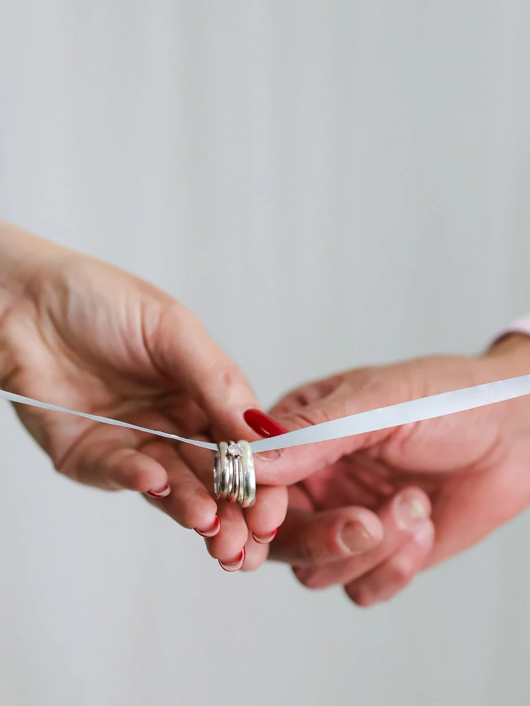 Close-up of two hands holding a wedding ring and a ribbon, with a background of a plain wall.