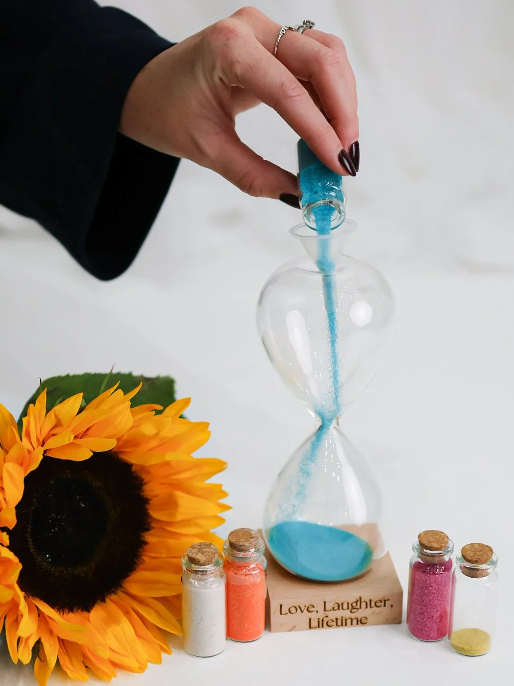 A person with dark nail polish is pouring blue sand into an hourglass-shaped glass vessel. There are four small jars of colorful powders and a large sunflower on a white surface. A wooden block with the inscription 'Love, Laughter, Lifetime' is also visible.