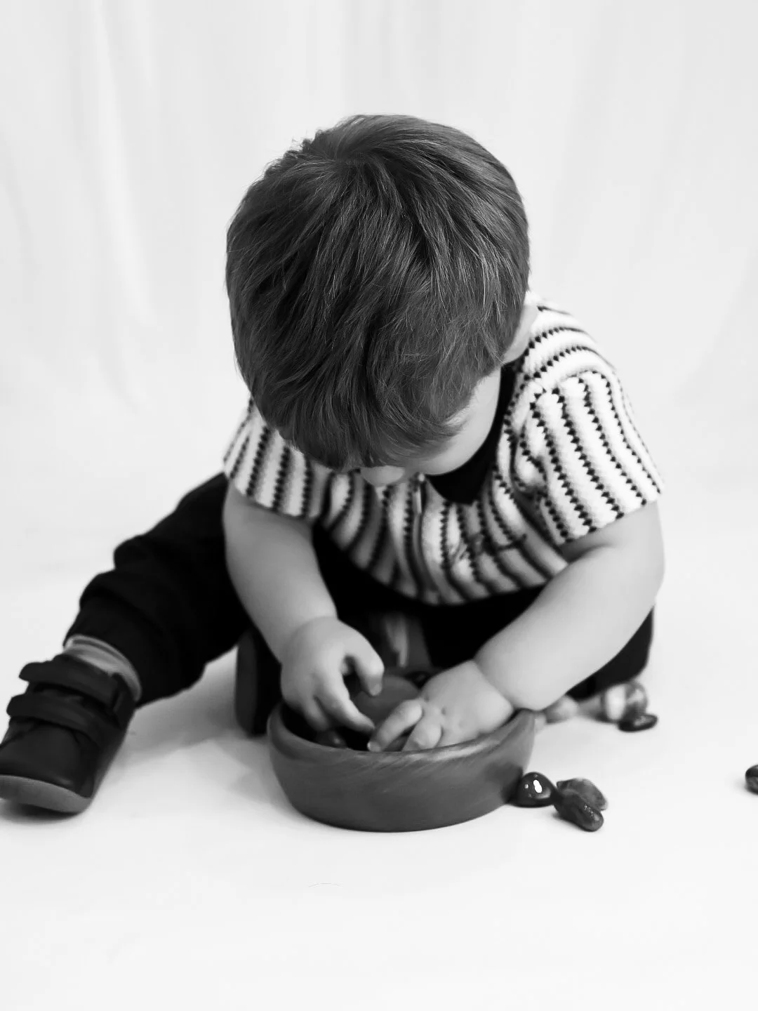 A young child with short hair, wearing a striped shirt, sitting on the floor, playing with stones inside a round bowl, with a few stones scattered around.