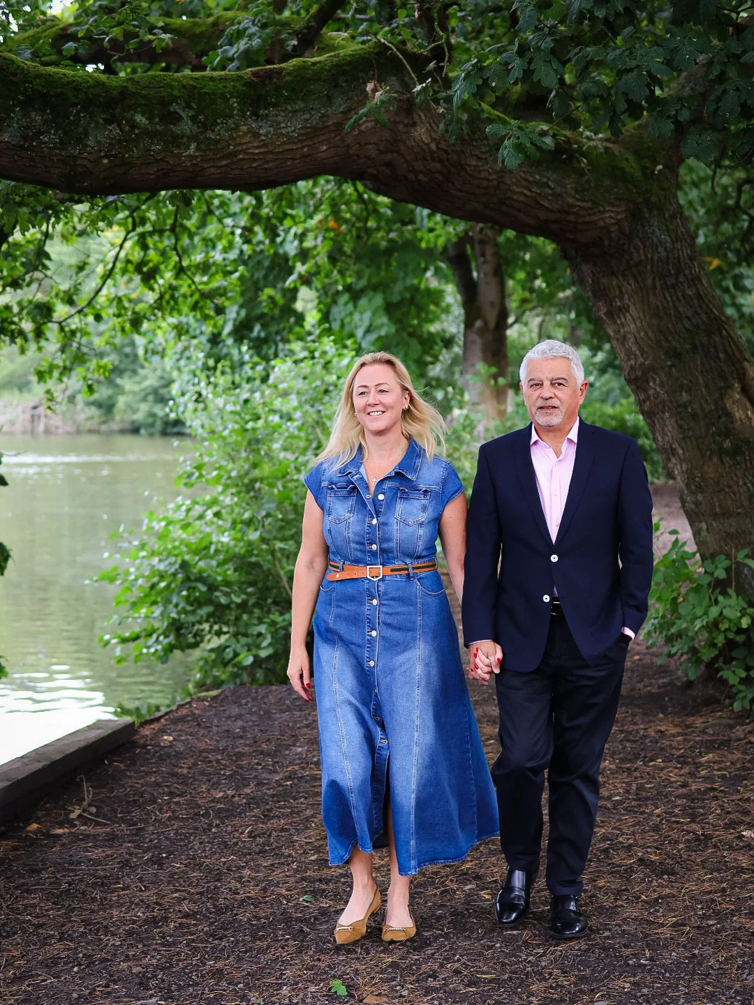A woman and a man walking hand in hand outdoors near a river, surrounded by lush green trees and foliage.
