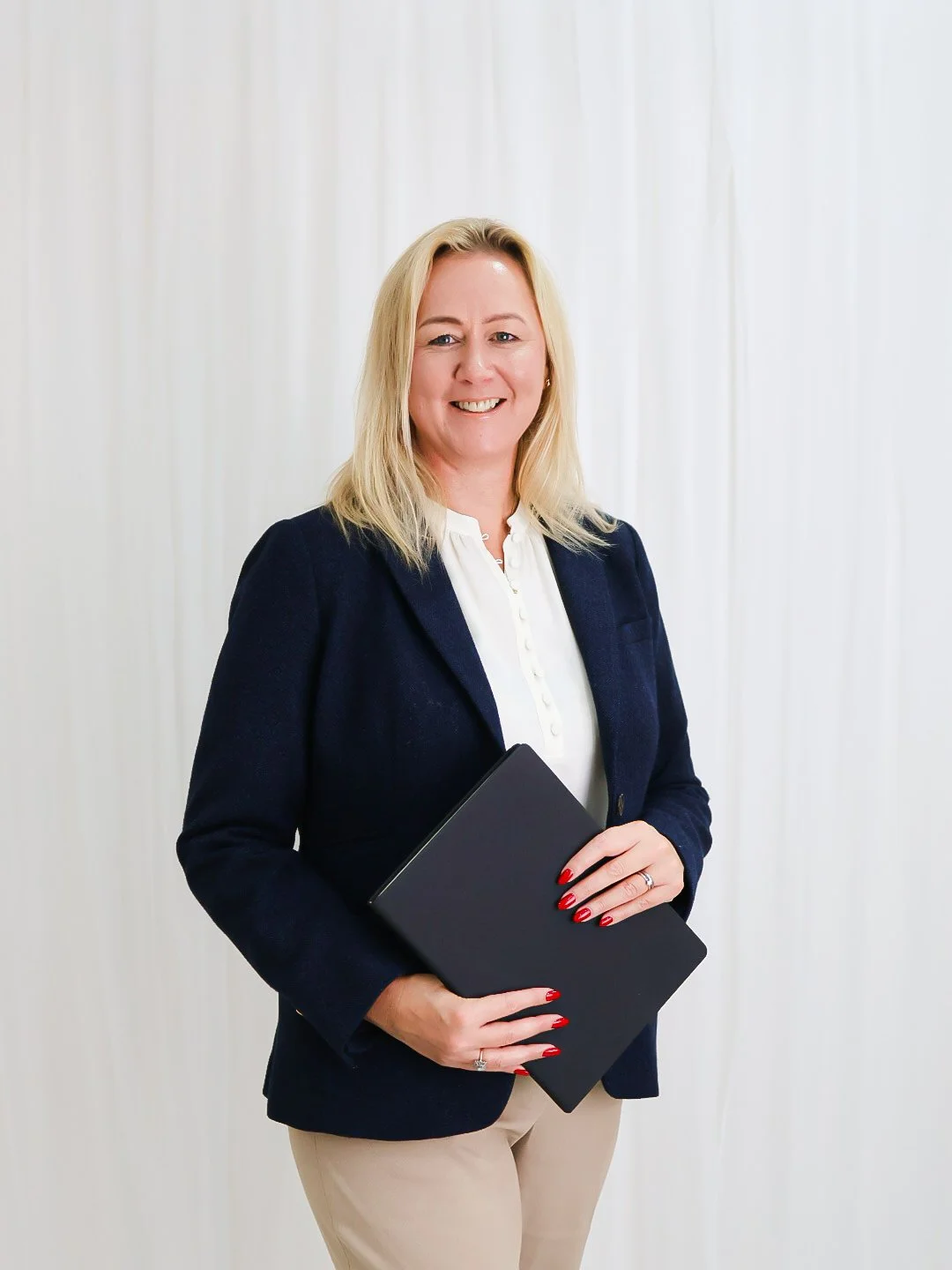 A professional blonde woman smiling, holding a closed black laptop, wearing a navy blazer, white blouse, and beige pants, standing in front of a white curtain background.