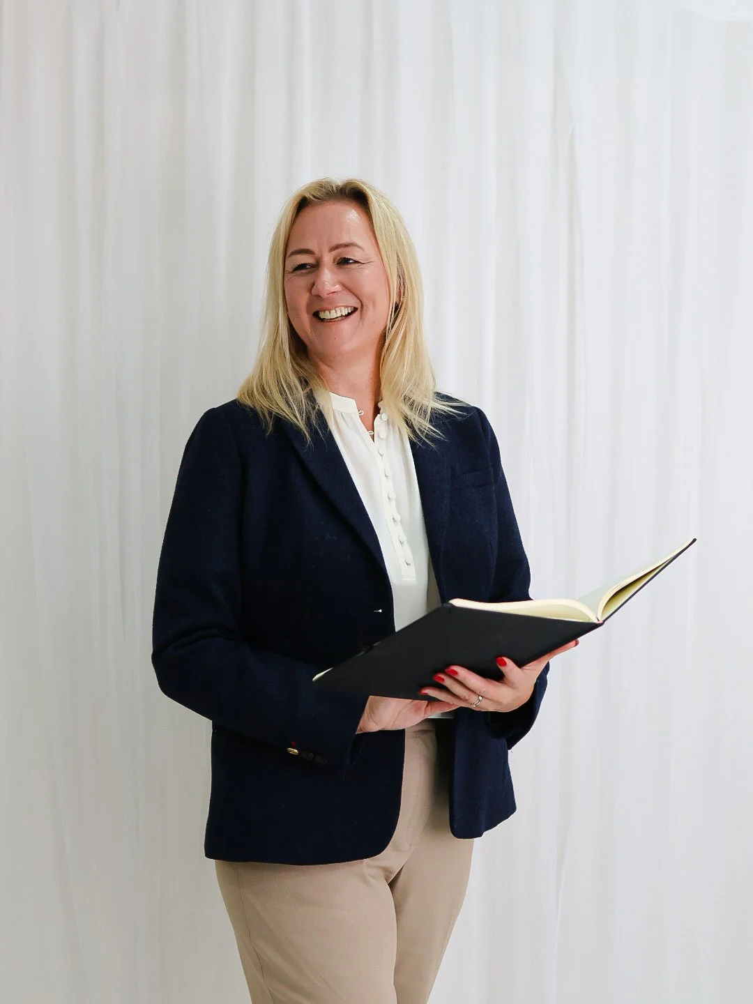A smiling woman with blonde hair, wearing a navy blazer and beige pants, holding an open notebook, standing against a white curtain background.
