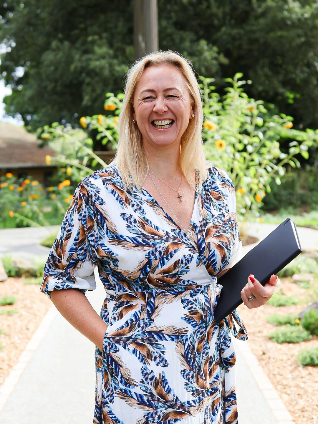 A woman in a floral dress smiling outdoors, holding a black folder in her right hand, with greenery and yellow flowers in the background.