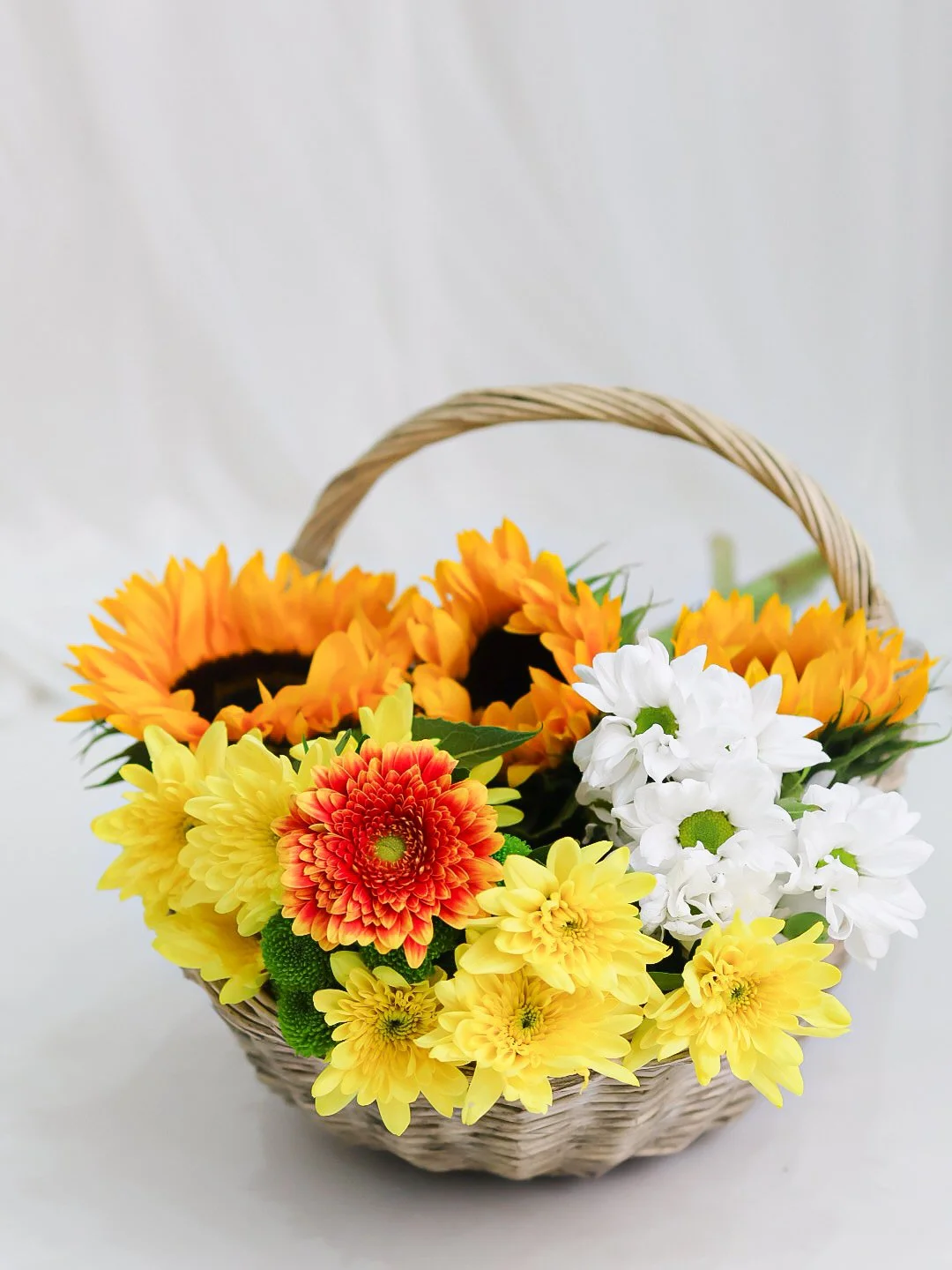 Basket filled with yellow, orange, red, and white flowers, including sunflowers and daisies, against a plain background.