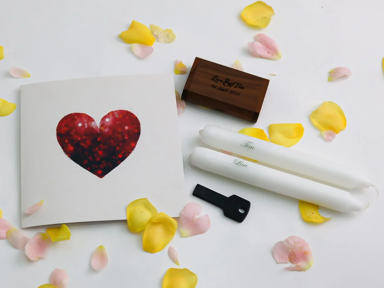 Wedding favors including a card with a red bokeh heart, three white rolled candles engraved with names, a small black key, a wooden box and scattered pink and yellow rose petals on a white surface.