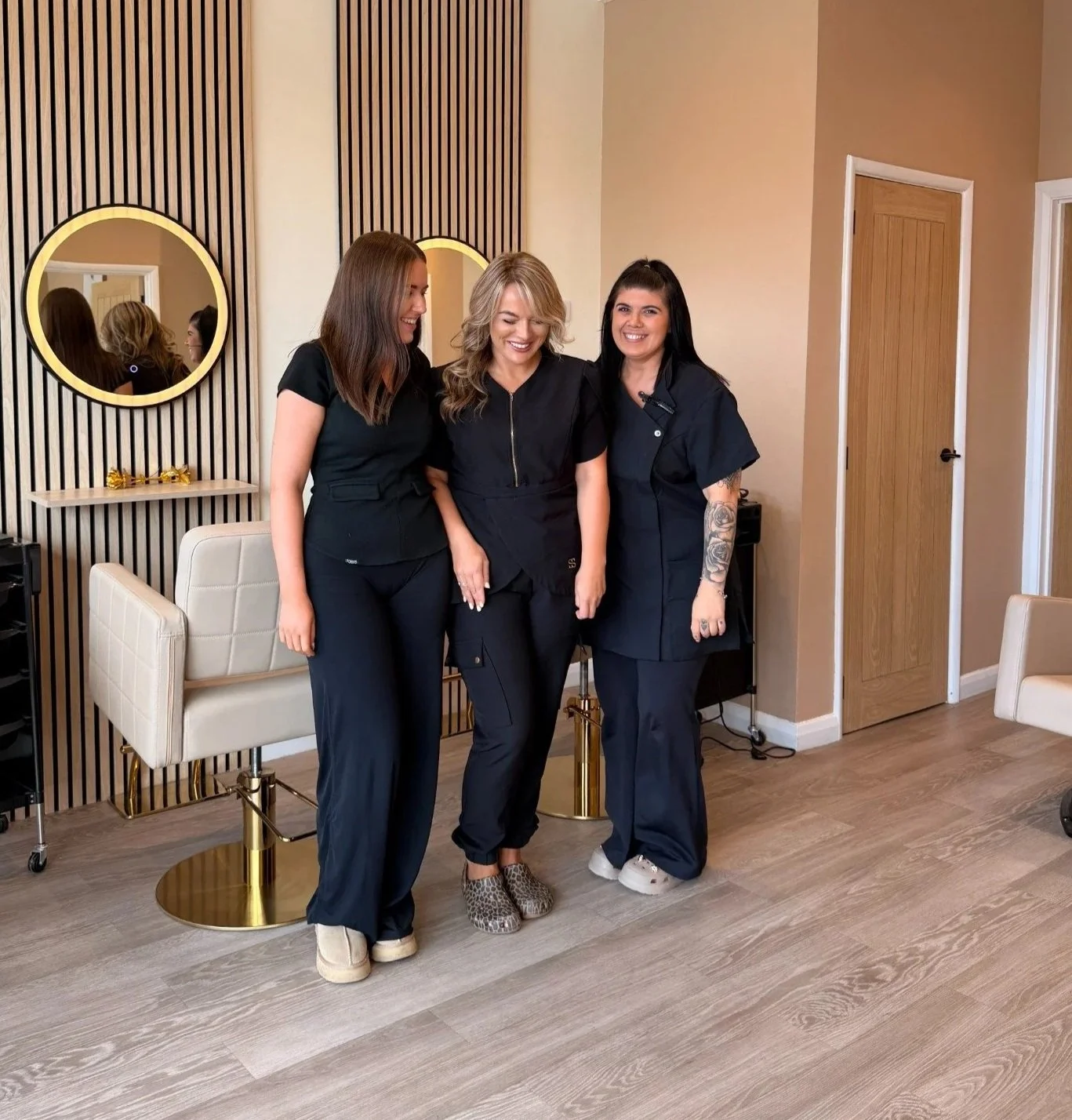 Three women in black scrubs standing together in a room with beige walls and wooden floors, smiling and posing for the photo.
