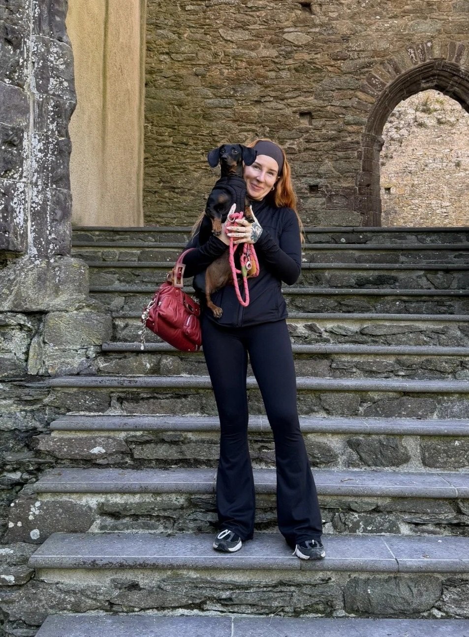 A woman with red hair holding a small black and tan dog on stone steps near an old stone building with a rounded arch window.
