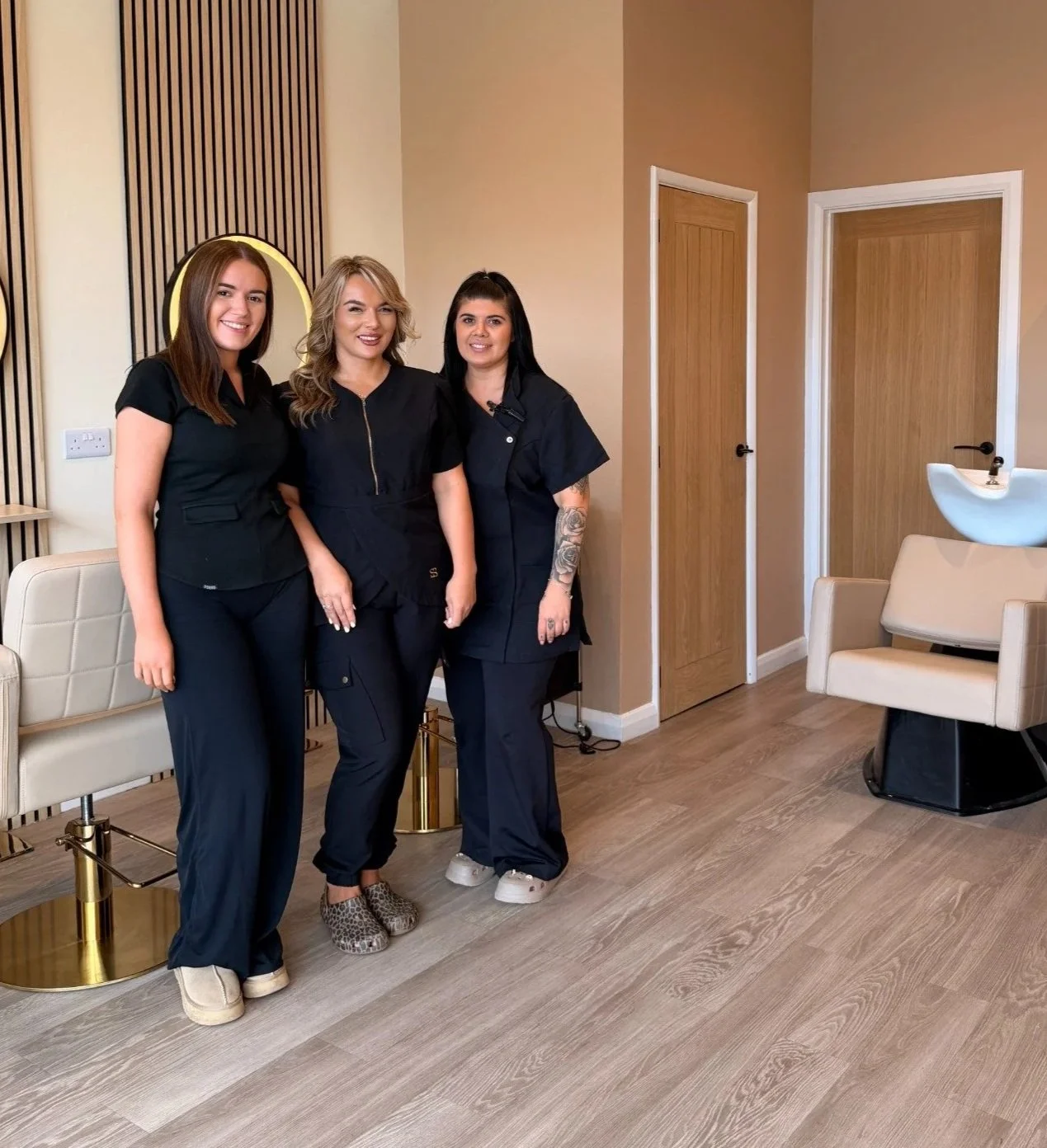 Three women standing together in a salon or spa setting, smiling at the camera, dressed in dark uniforms, with salon chairs, a sink, and closed doors in the background.