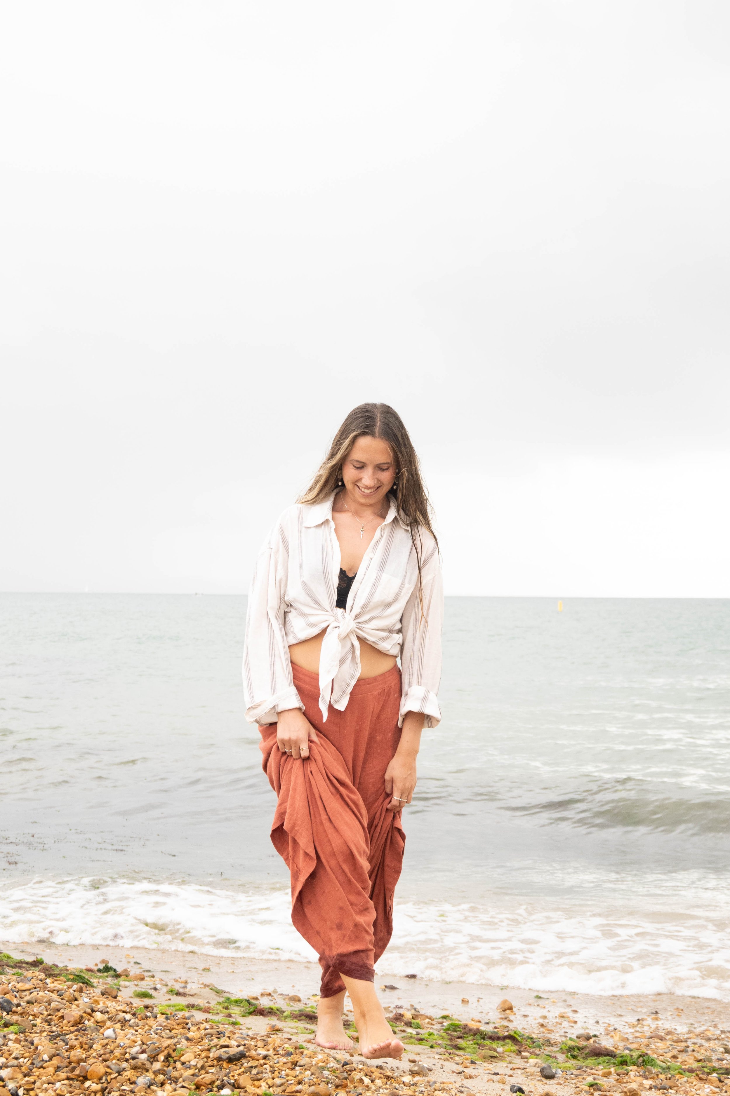 A woman walking barefoot on a pebble beach near the water, smiling and looking down, with overcast sky in the background.