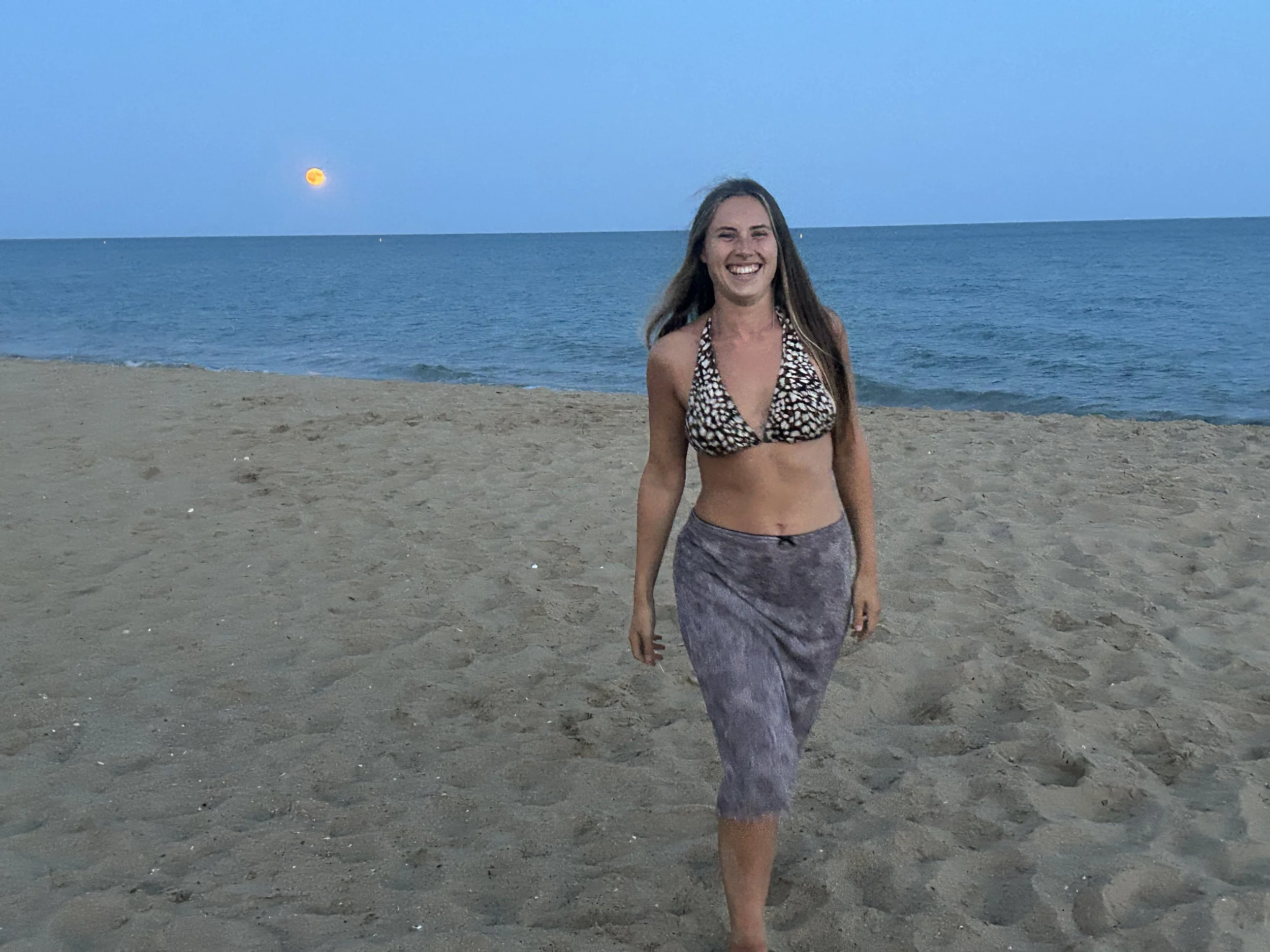 A woman with long hair smiling on a sandy beach during dusk, wearing a patterned bikini top and gray beach pants, with the ocean and a setting moon in the background.