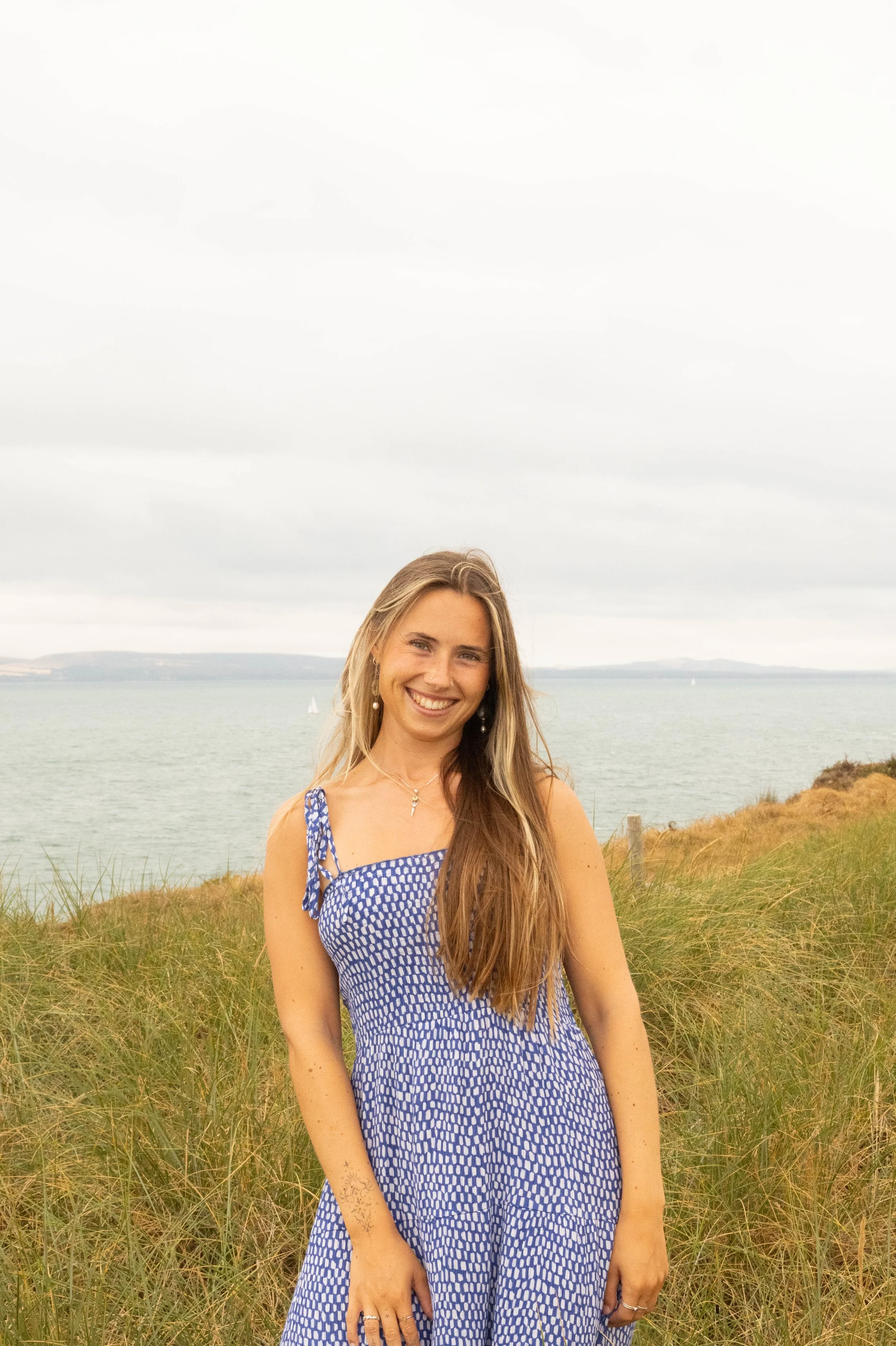 A young woman with long light brown hair wearing a blue and white patterned sundress, standing in a grassy area near a body of water, with hills and sailboats in the background under a cloudy sky.