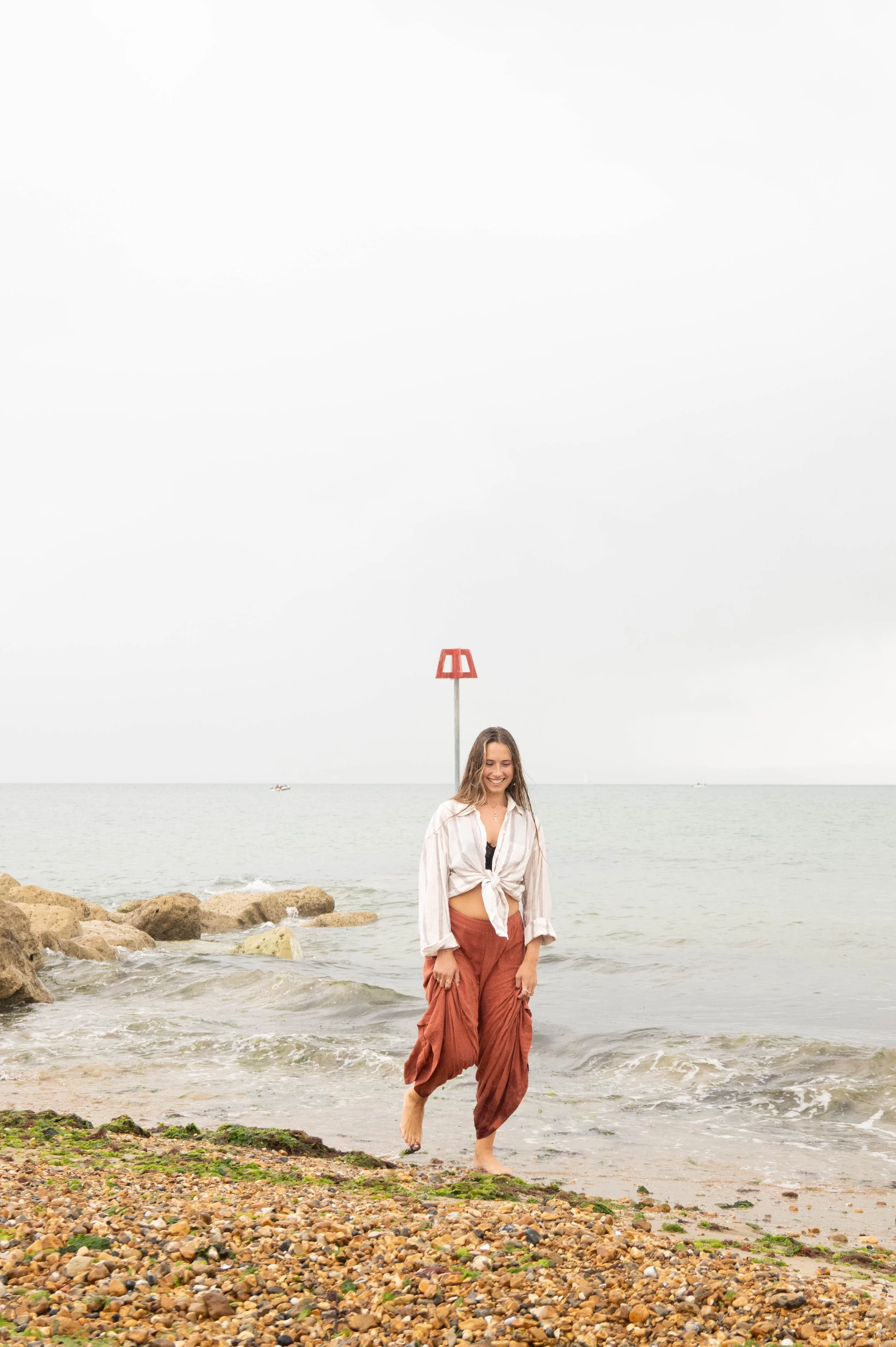 A woman with long hair, wearing a tied white blouse and loose brown pants, walking barefoot on rocks and pebbles at a beach with overcast sky and calm sea, with a small boat and a red and white tower in the background.