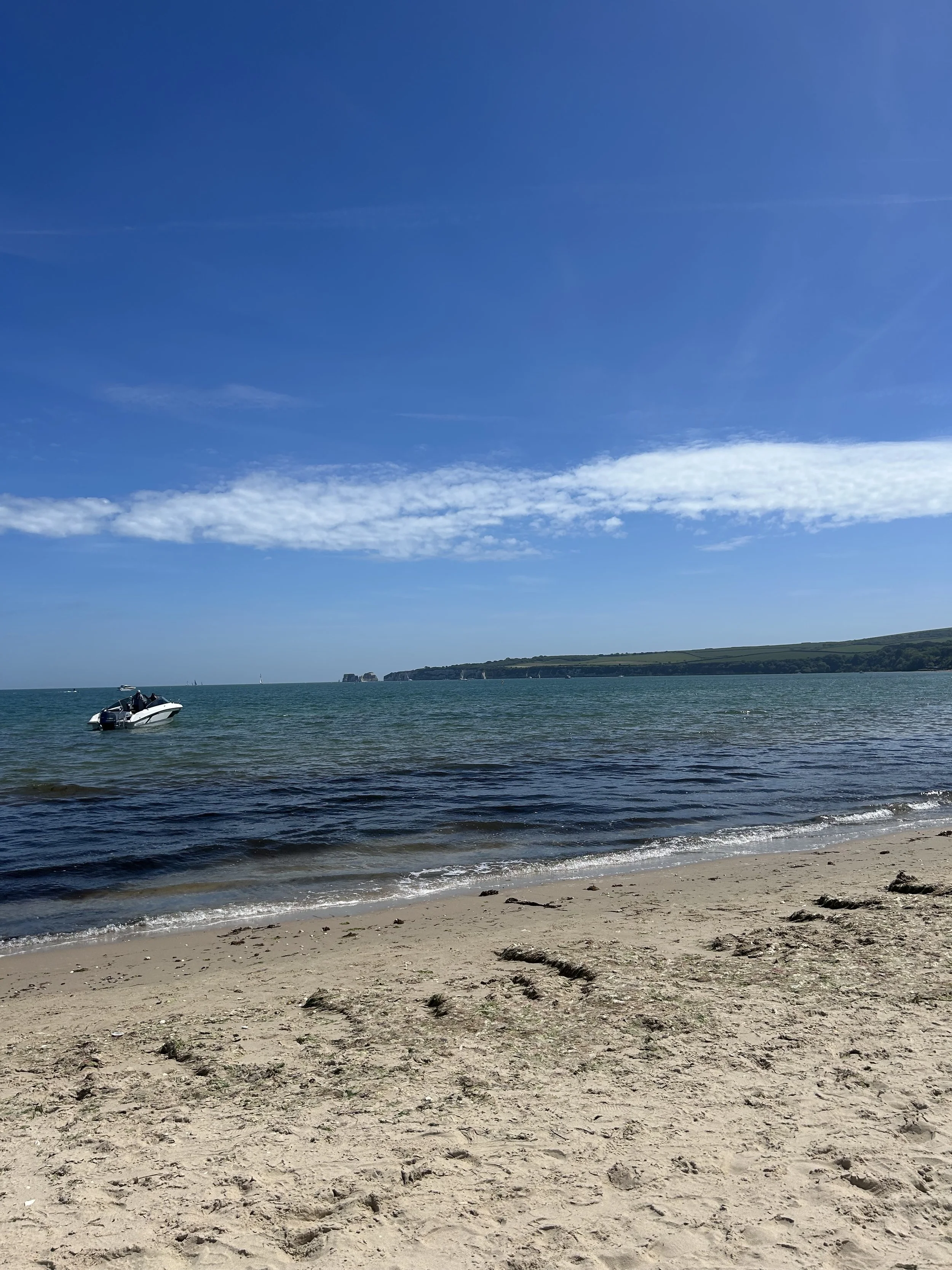 A sandy beach with small rocks and footprints, calm ocean water with a few boats, and a clear blue sky with some clouds and distant land on the horizon.