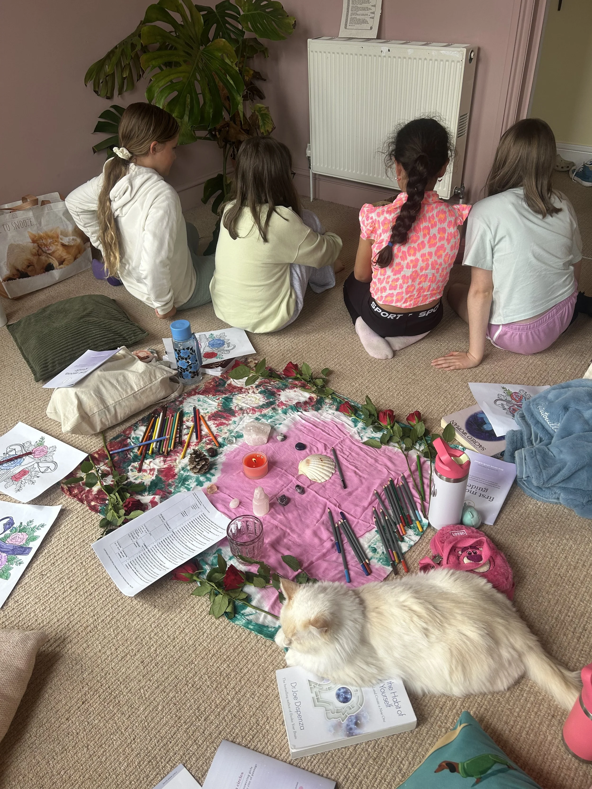 Group of four children sitting on the carpeted floor engaged in arts and crafts, with coloring pages, colored pencils, decoration items, a book titled 'The History of Alzheimer’s Disease' by Dr. Joe Dispenza, and a large white dog lying nearby, in a room with a pink wall and a large leafy plant.