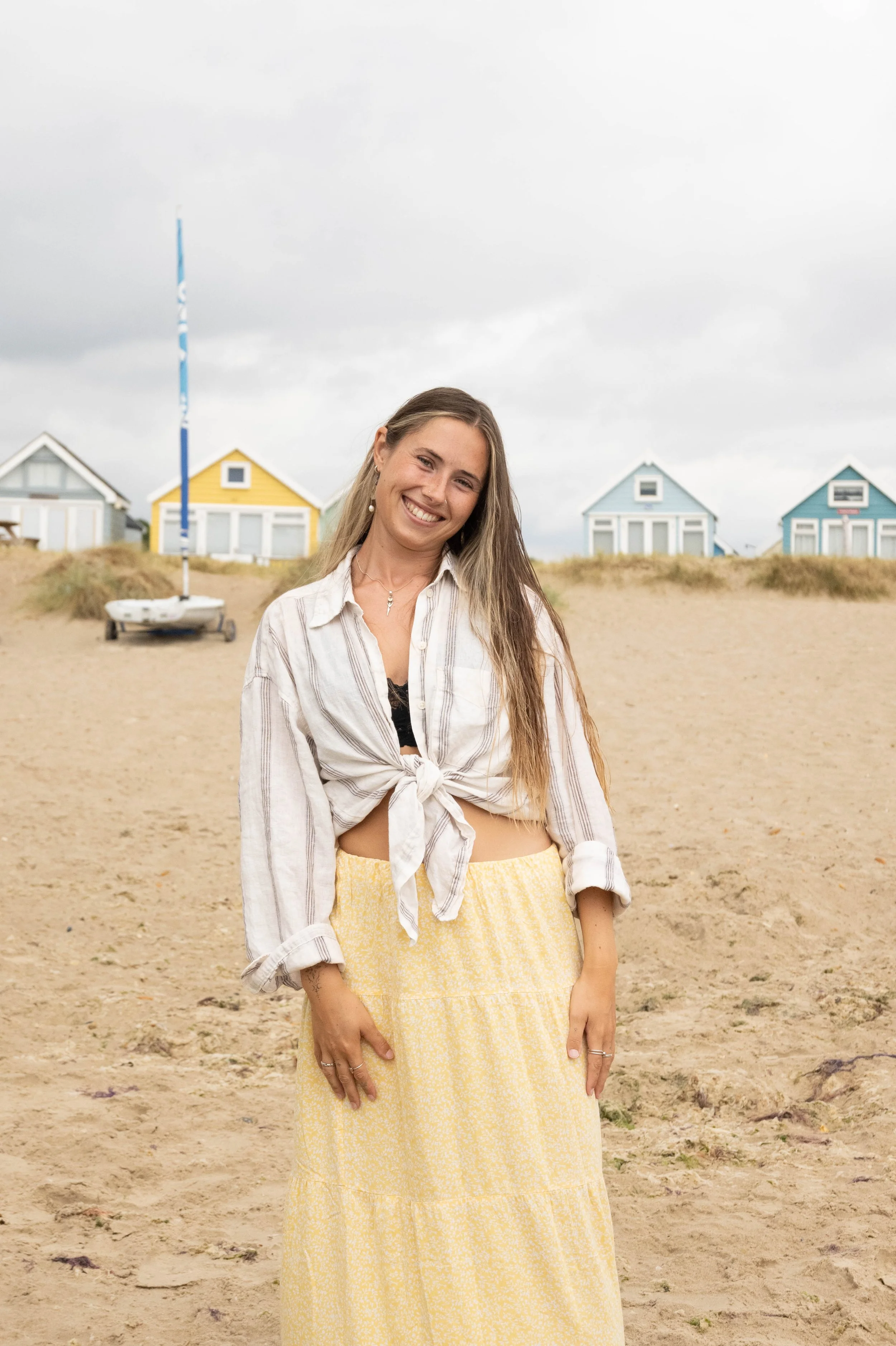 A woman with long hair smiling on the beach, wearing a white shirt tied at the waist and a yellow skirt, with colorful small houses and a sailboat in the background under cloudy skies.