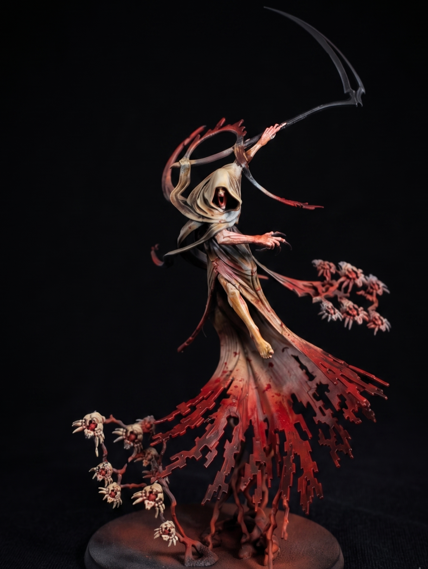 Miniature figurine of a skeletal figure with a hood, holding a scythe, standing amidst red, leafless trees and smaller skull-like structures on a round base.