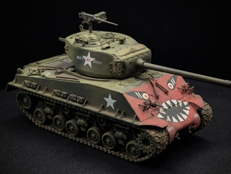 Model of a military tank with a cartoonish face painted on the front, featuring sharp teeth and eyes, painted in red and green with white star insignias, and the words "Mice's Red Devils" on the side, placed on a black surface against a black backgro