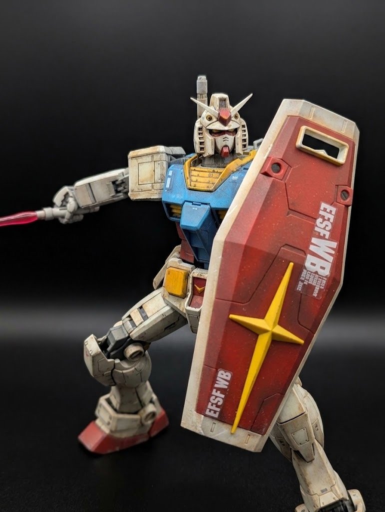 A detailed model of a Gundam robot with a red and yellow shield, holding a weapon, set against a dark background.