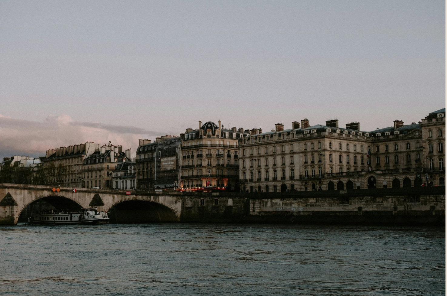 Historic Paris buildings along the Seine River at dusk, with a stone bridge and boat