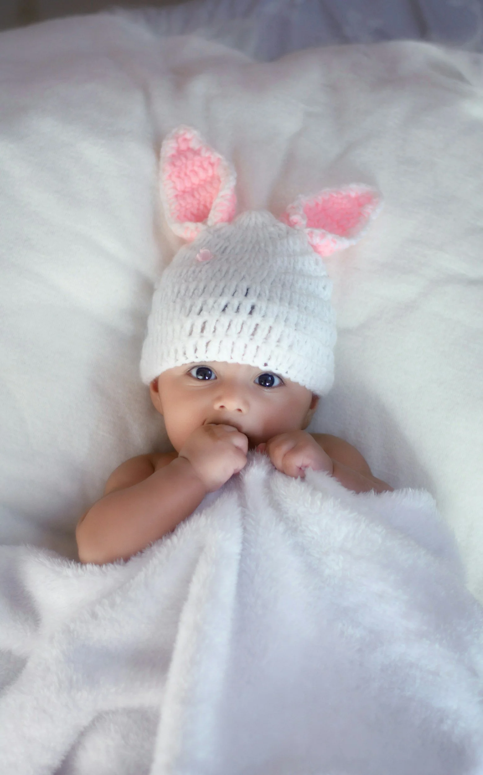 A baby with big blue eyes, wearing a white knit hat with pink ears, lying on a soft white blanket.
