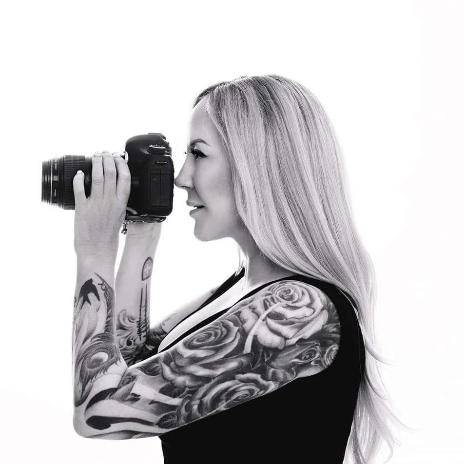 A woman with long, blonde hair and tattoos on her arms, holding a camera up to her face, taking a photo.