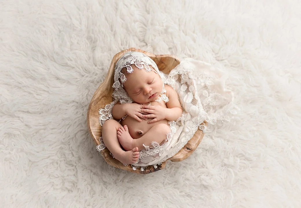 Sleeping baby wrapped in lace, lying in a wooden bowl on a fluffy white surface.