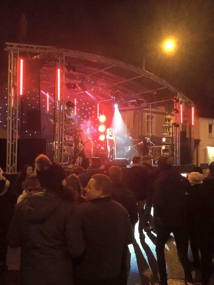 A nighttime outdoor concert with a live band performing on a stage illuminated with red and white lights, crowd gathered in front, some people taking photos, and city buildings in the background.