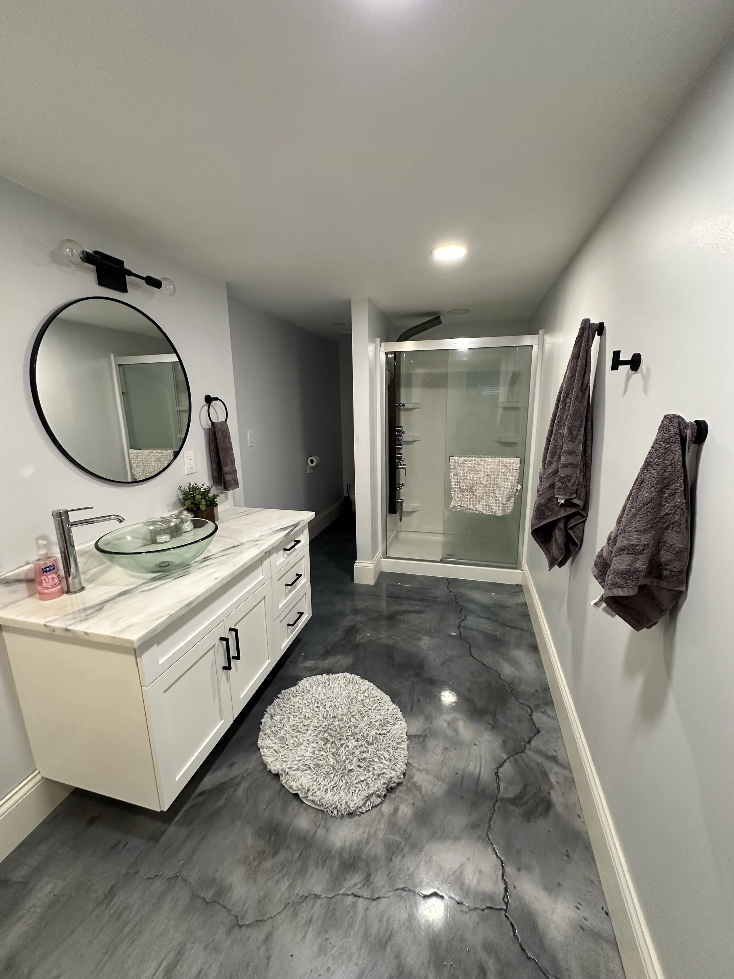 A modern bathroom with a white vanity, a circular mirror, black fixtures, towels hanging on black hooks, a shower with clear glass door, and a textured white rug on dark marble flooring.