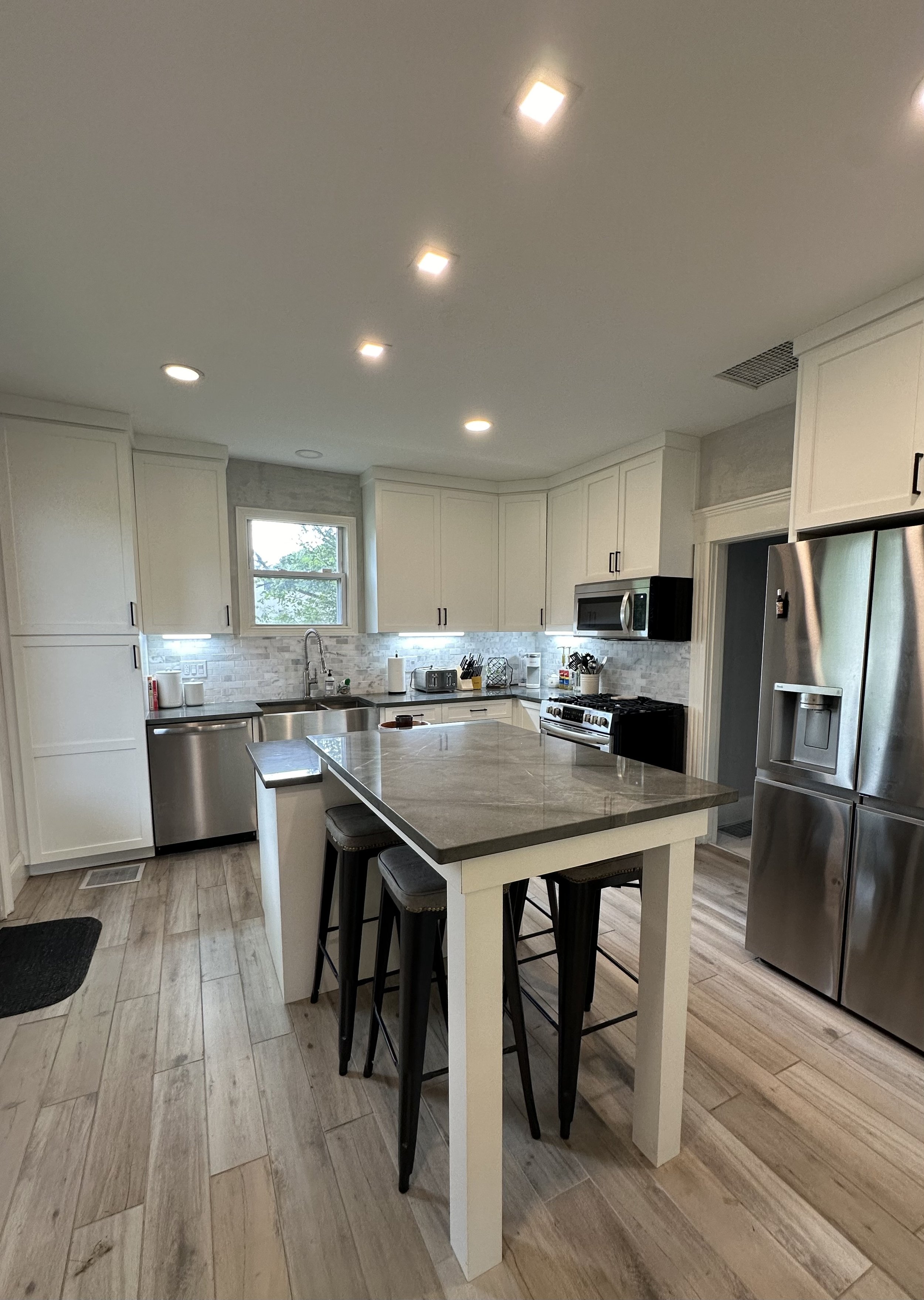 Modern kitchen with white cabinets, stainless steel appliances, a central island with a marble countertop, a window above the sink, and wood-look flooring.