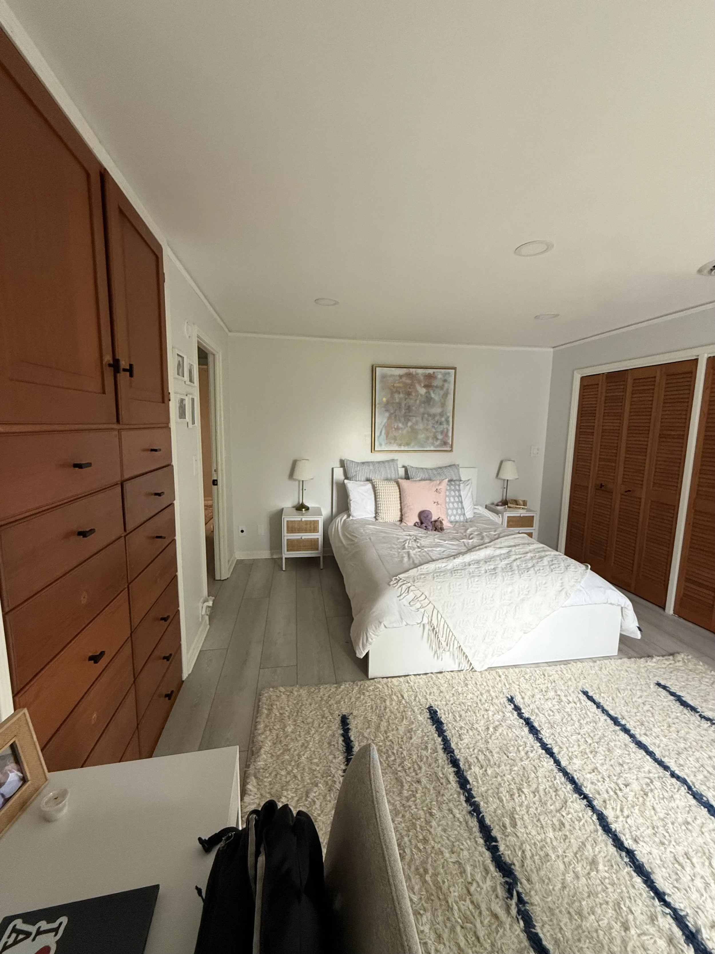 The image shows a cozy bedroom with a bed covered in white bedding and decorated with various pillows. On each side of the bed, there are matching nightstands with lamps. A framed artwork hangs above the bed. To the left, there are large wooden cabin