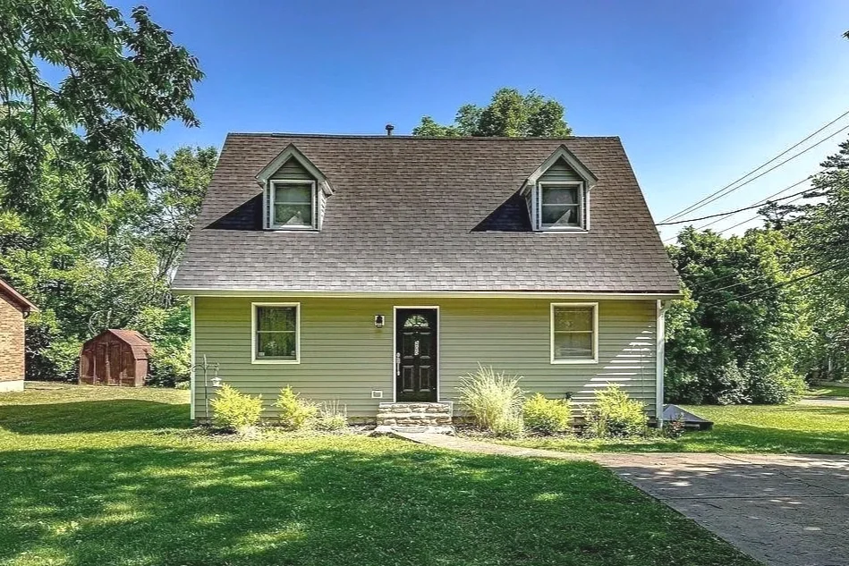 A green two-story house with dormer windows on a sunny day, surrounded by a lawn and trees.