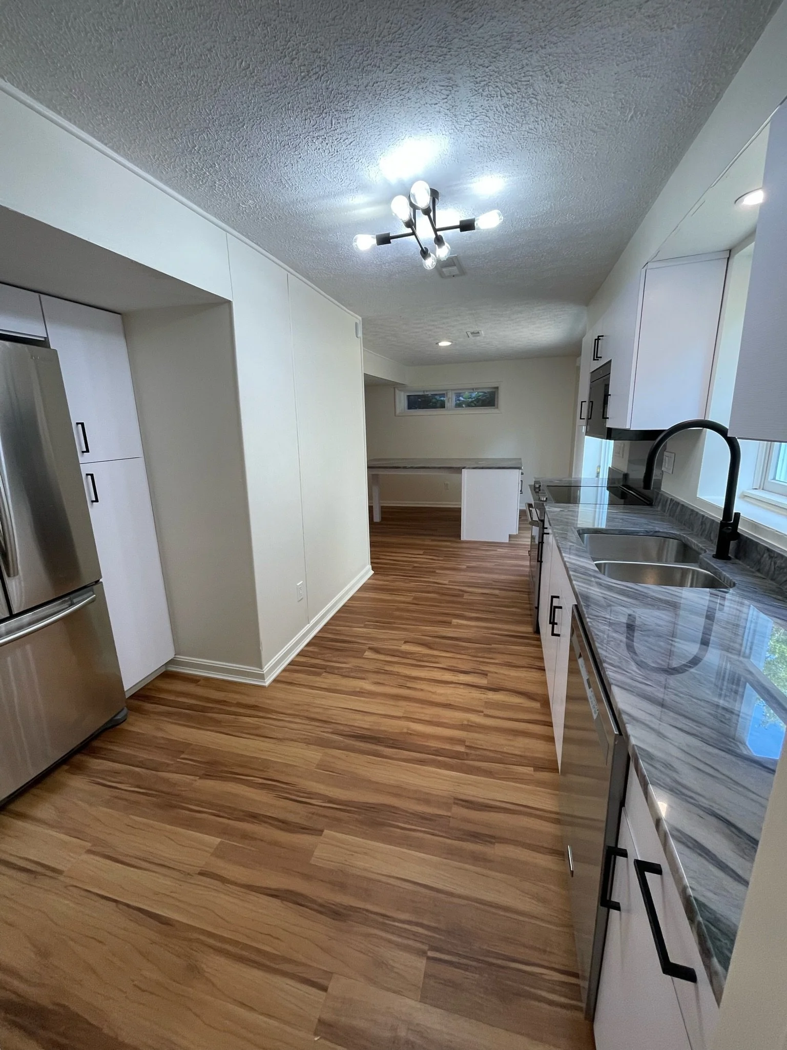 Modern kitchen with white cabinets, marble countertops, stainless steel appliances, double sink, black faucet, hardwood flooring, and a ceiling light fixture.