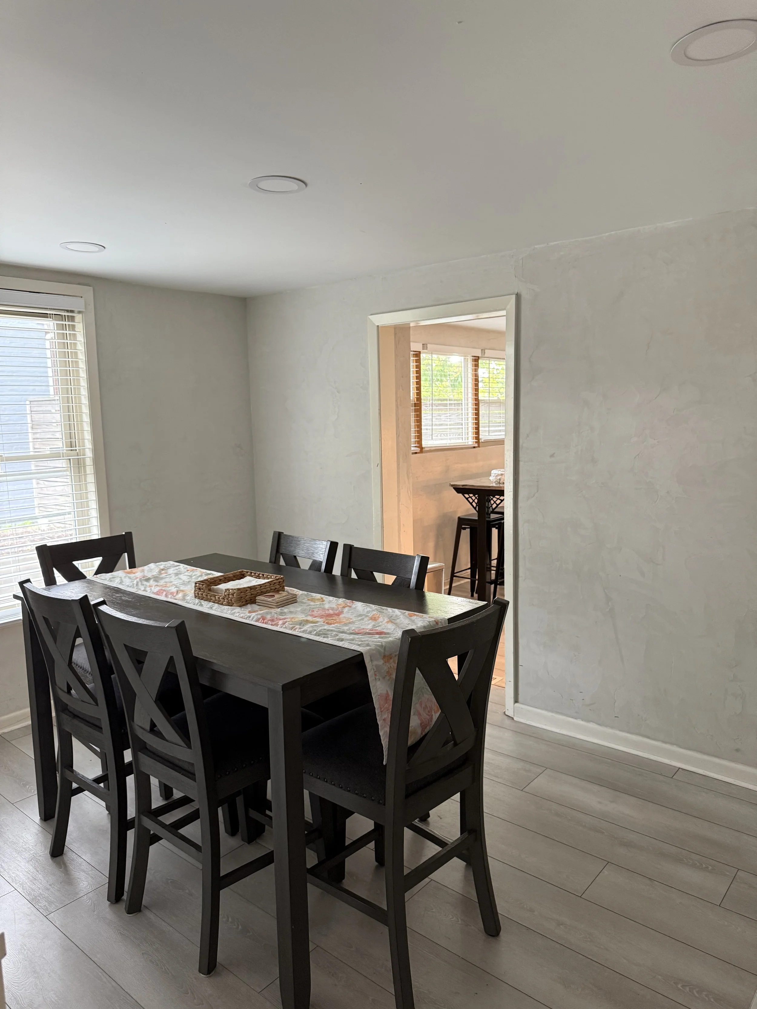 A dining room with a black wooden table and matching chairs, a window with blinds, and a doorway leading to a sunlit room with a high table and chairs.