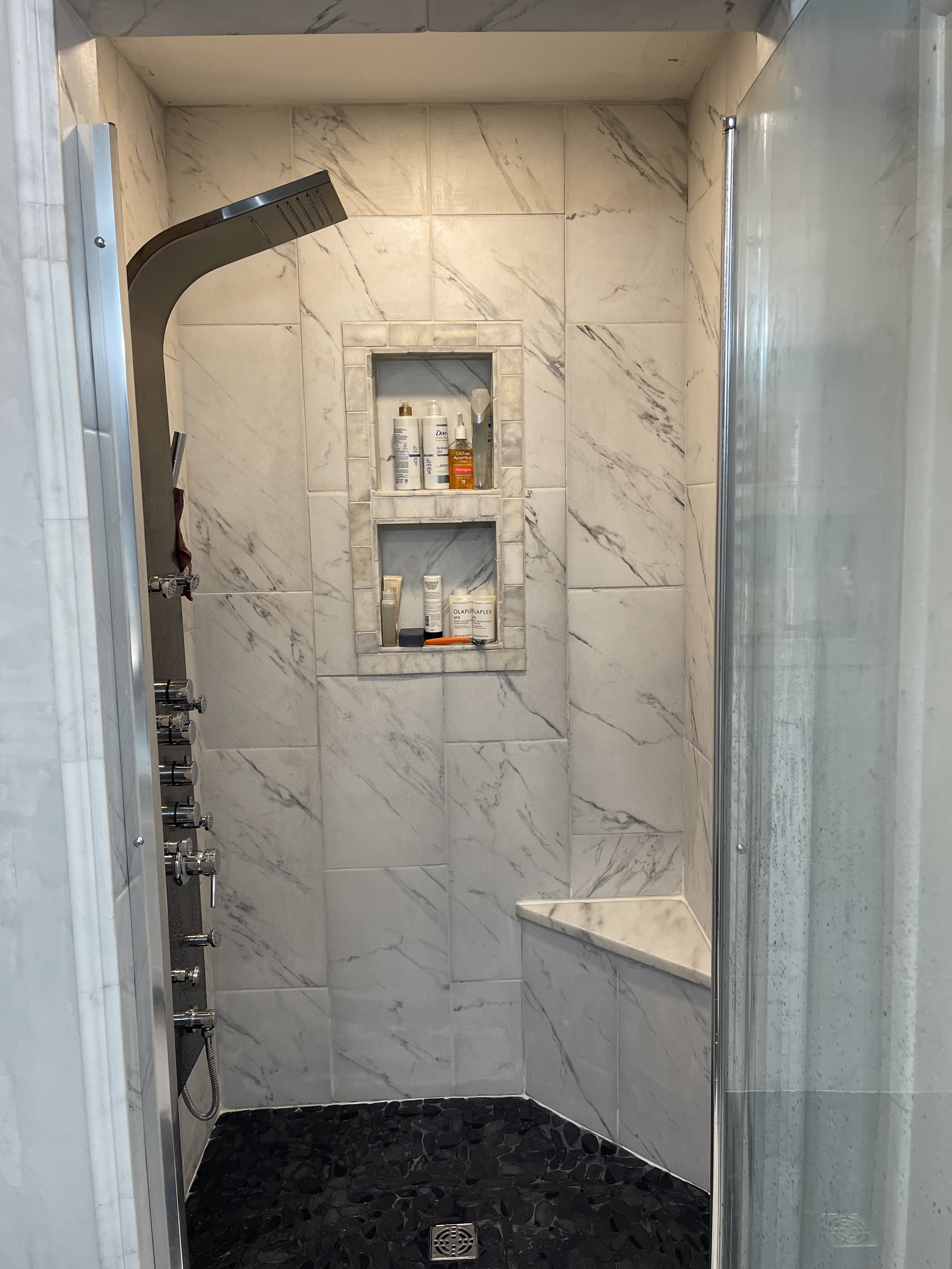A modern shower with marble tiles on the walls, a built-in shelf with toiletries, a black pebble stone floor, and a glass door.